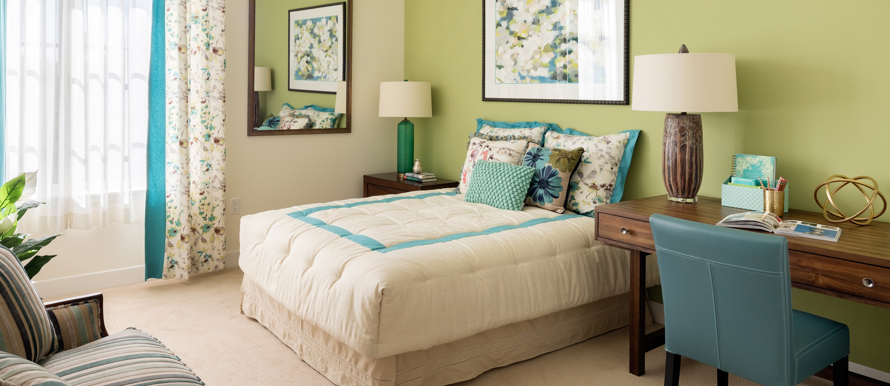 A cozy bedroom with a neatly made bed featuring floral and turquoise accents, a wooden desk with a lamp and books, a teal chair, artwork on green walls, and a window with sheer floral curtains letting in natural light.