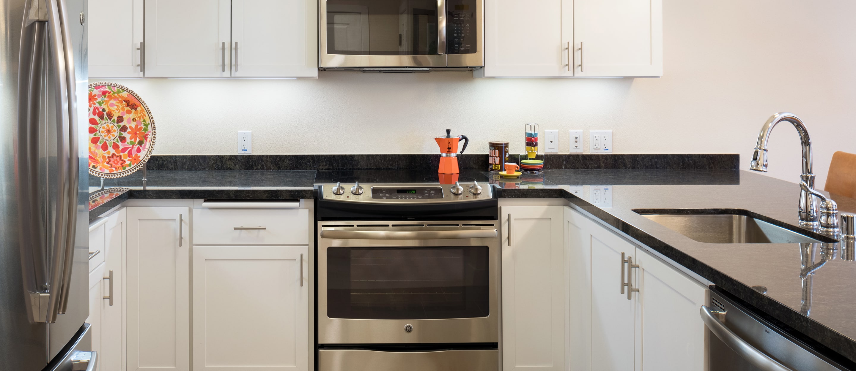 Modern kitchen with white cabinets, black countertops, stainless steel appliances, a colorful plate on the left, and a red coffee maker on the stove. Double sink and stacks of bowls are on the right.