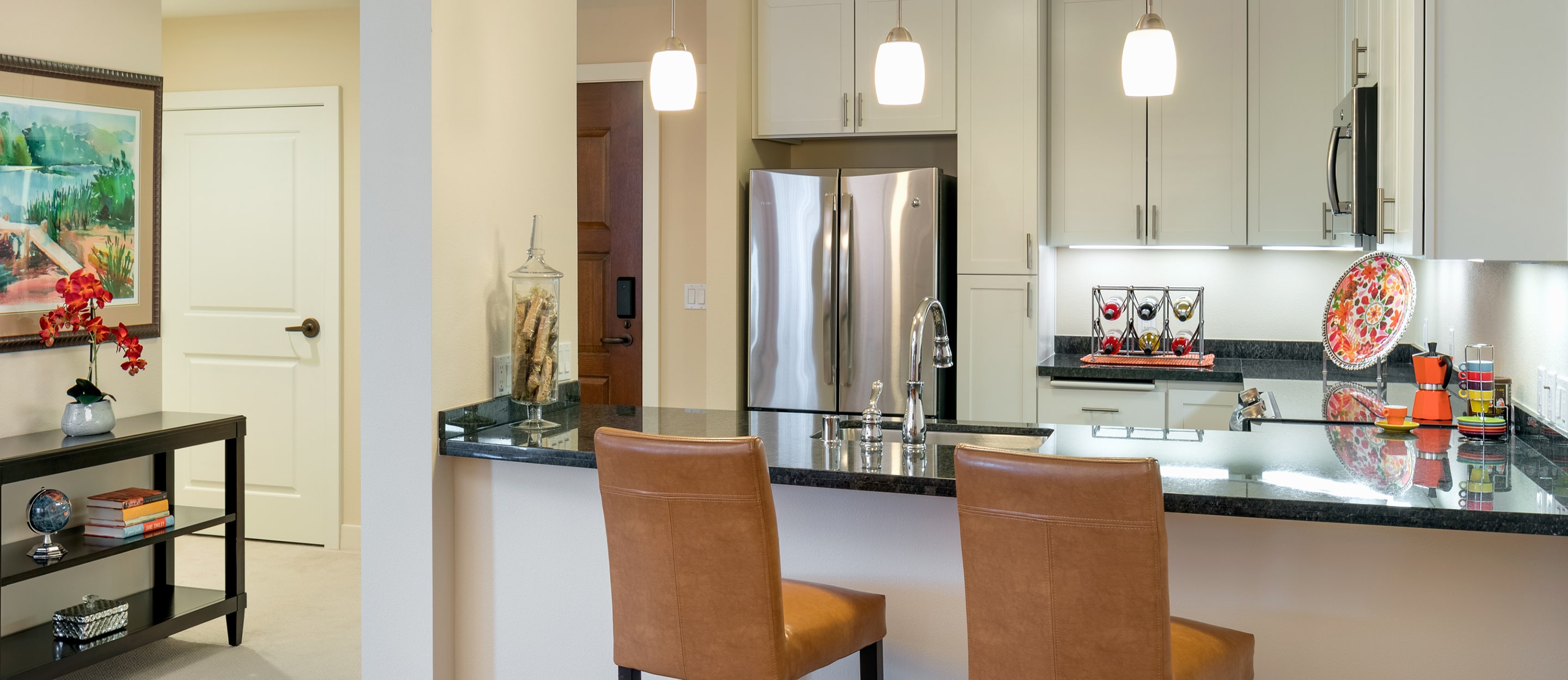 Modern kitchen with white cabinets, stainless steel appliances, black granite countertops, and two brown leather bar stools. Pendant lights hang overhead, and colorful decor accents the space. A small living area is visible to the left.