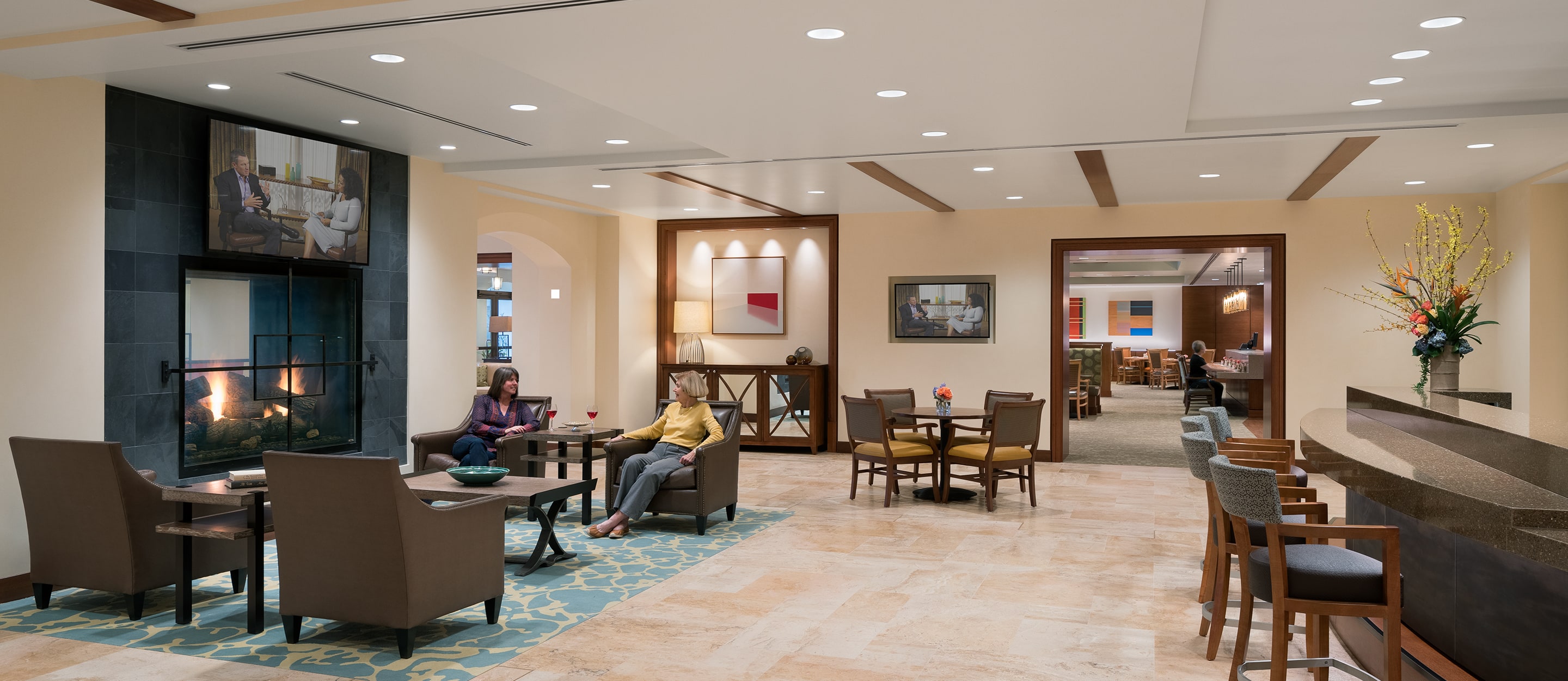 A spacious, modern lounge with armchairs and small tables, a fireplace, wall-mounted TV, and artwork. People are seated and talking. There’s a bar on the right and a bright, open dining area in the background.