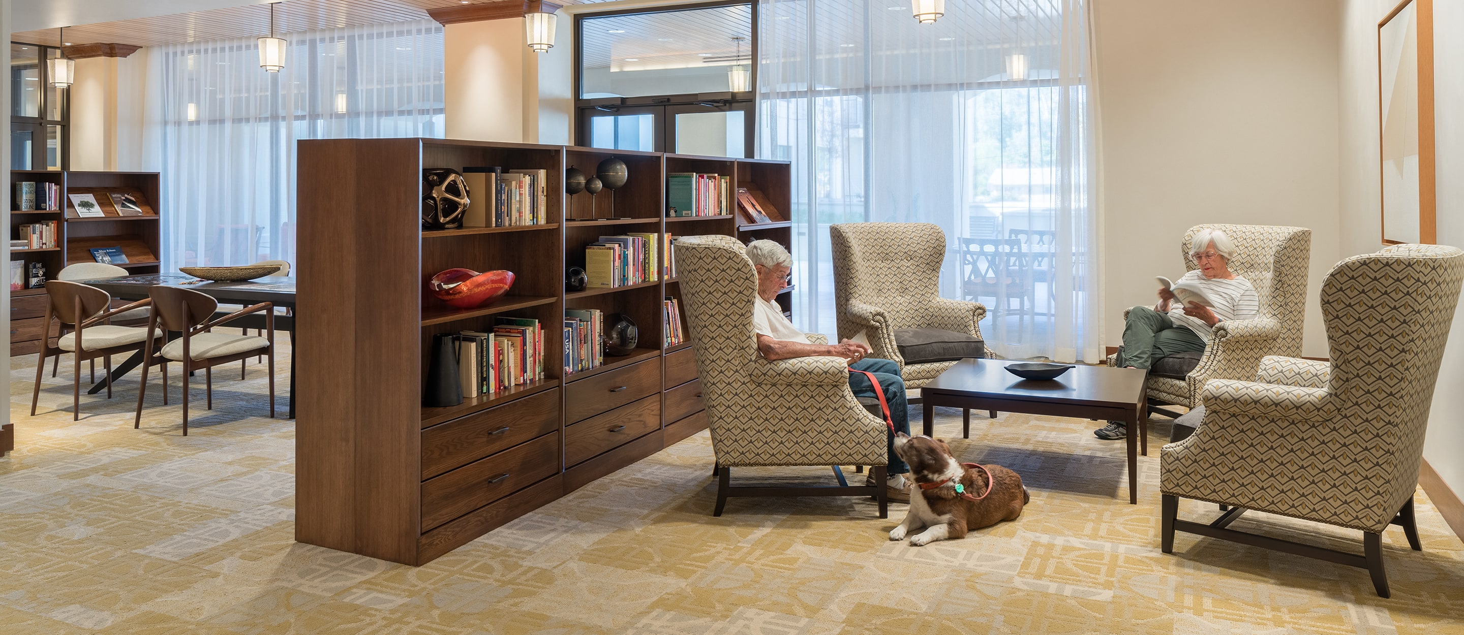 Two elderly people sit and read in armchairs in a cozy library or lounge area, divided by a wooden bookshelf. A dog rests on the carpet, and large windows let in natural light. Books and artwork decorate the space.