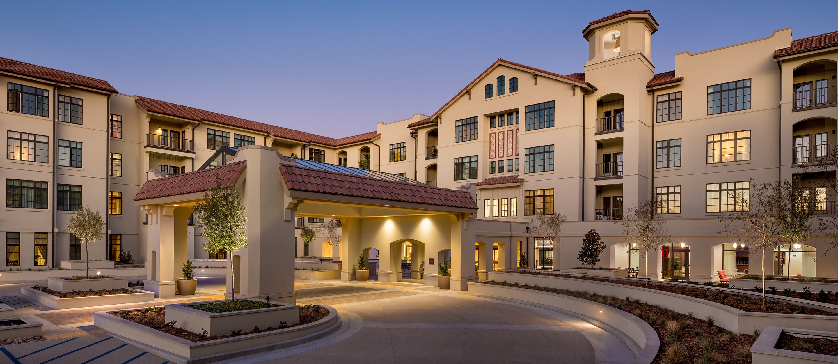 A modern Mediterranean-style apartment building with a red-tiled roof, arched entryway, and well-lit courtyard featuring landscaped flower beds and pathways at dusk.