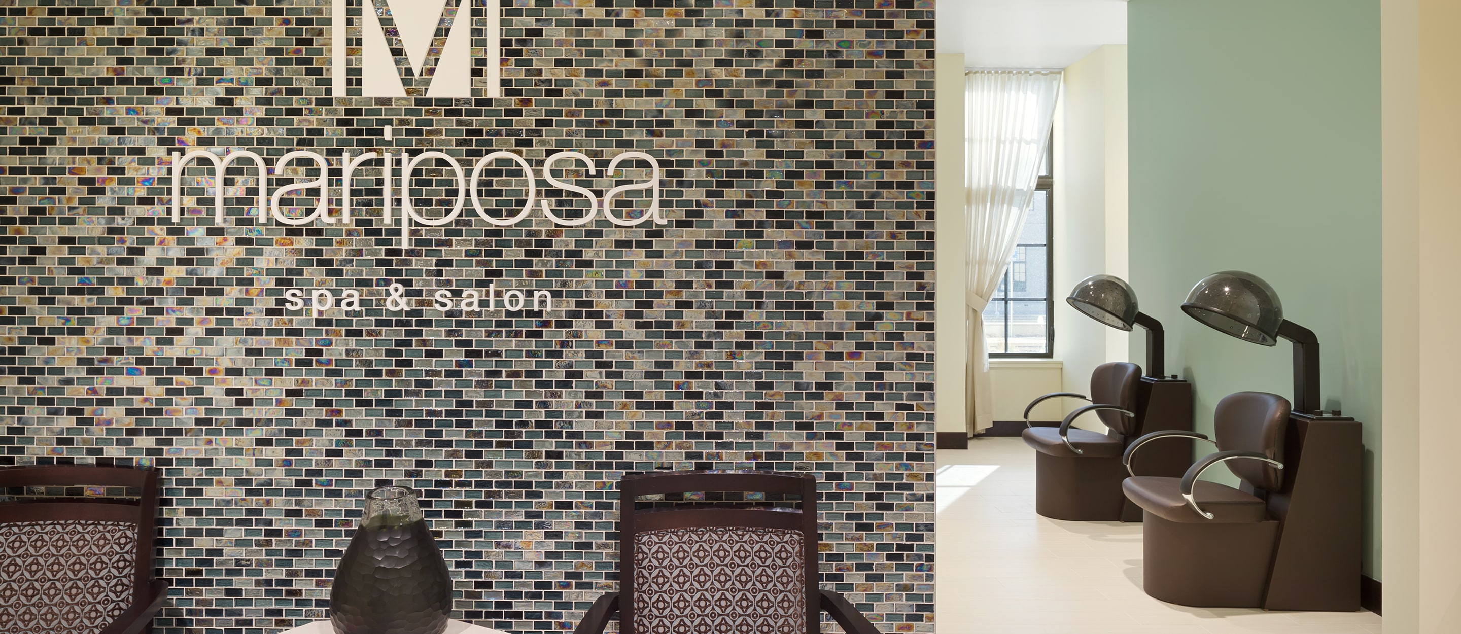 Reception area of Mariposa Spa & Salon with a tiled wall, modern chairs, and two black salon hair dryers in a brightly lit room with large windows and sheer curtains.