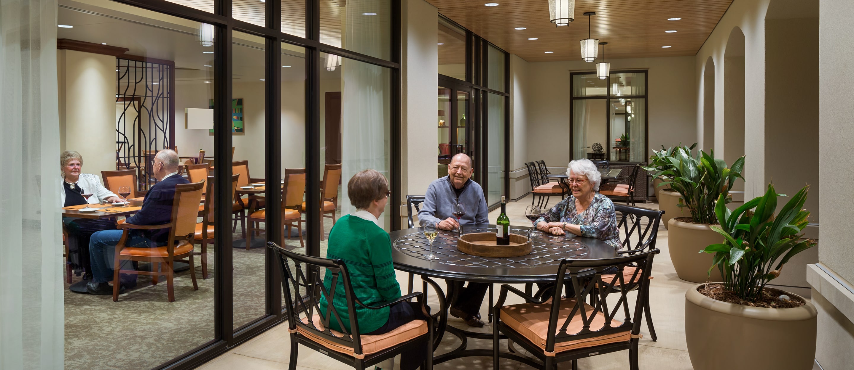 Four older adults sit and talk around an outdoor patio table, while two other older adults dine inside a brightly lit restaurant visible through large glass windows. The space is modern with plants and warm lighting.