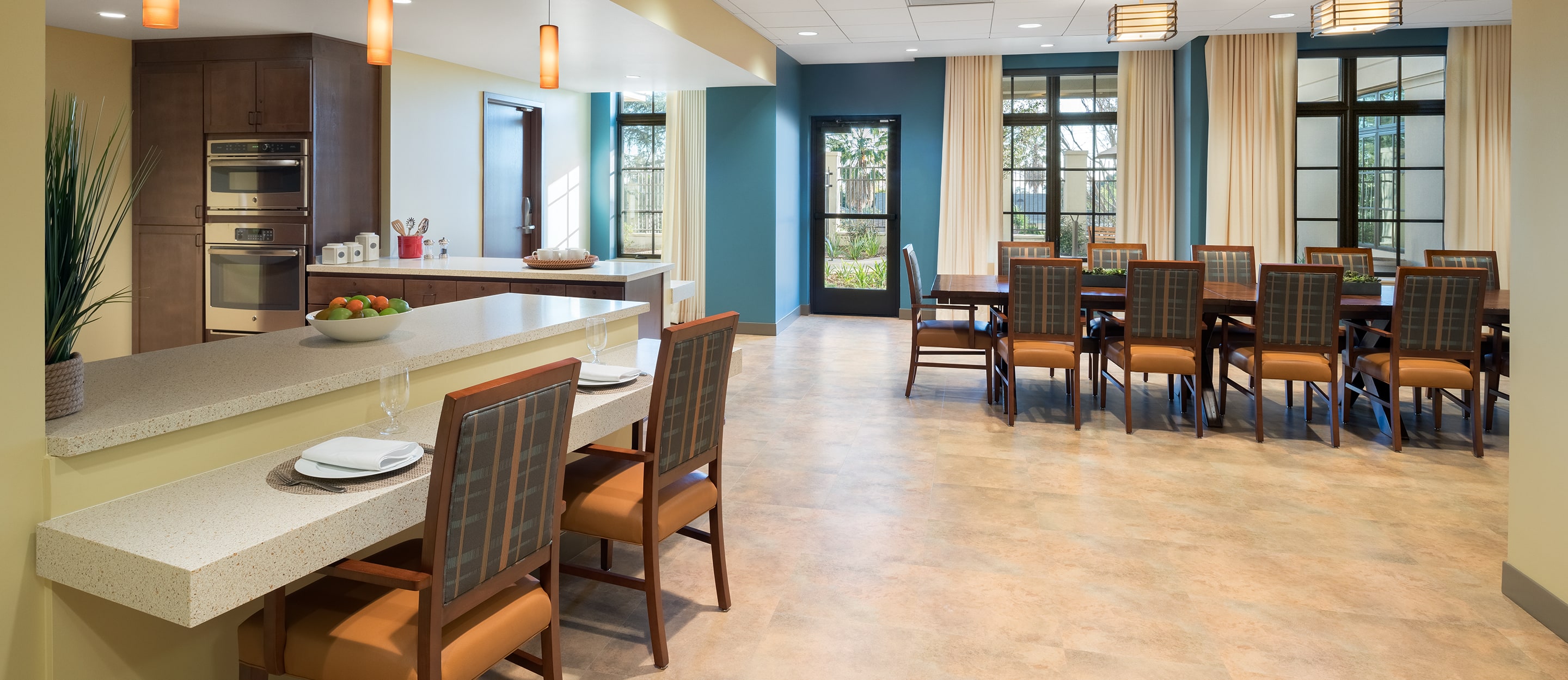 A spacious, modern dining area with two dining tables and several chairs, a kitchen with built-in appliances, a breakfast bar with seating, pendant lighting, and large windows letting in natural light.