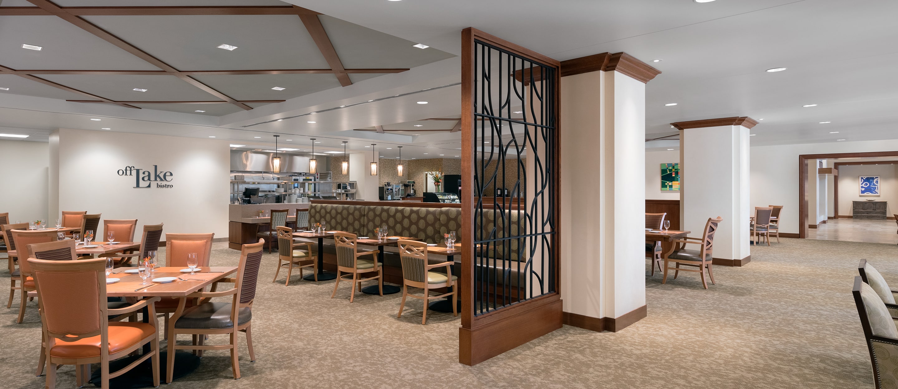 Spacious, modern restaurant interior with light brown chairs and tables, beige carpet, cushioned booths, open kitchen area, and contemporary decor. The space is well-lit with recessed lighting and wooden accents.