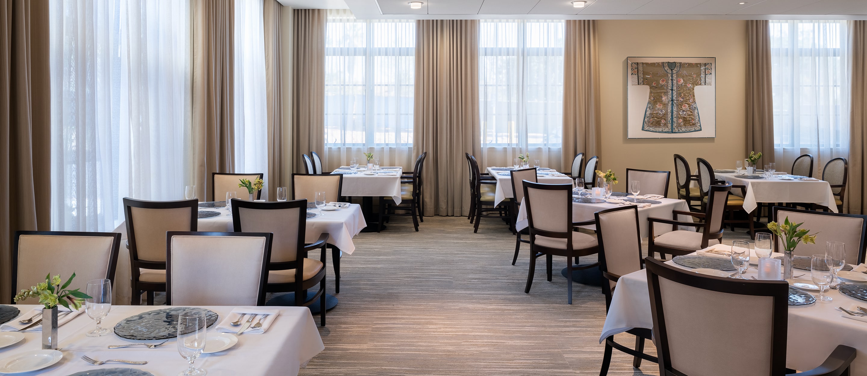 Elegant restaurant dining room with neatly set tables and cream-colored chairs, large windows with sheer curtains allowing natural light, and a decorative kimono artwork on the wall.