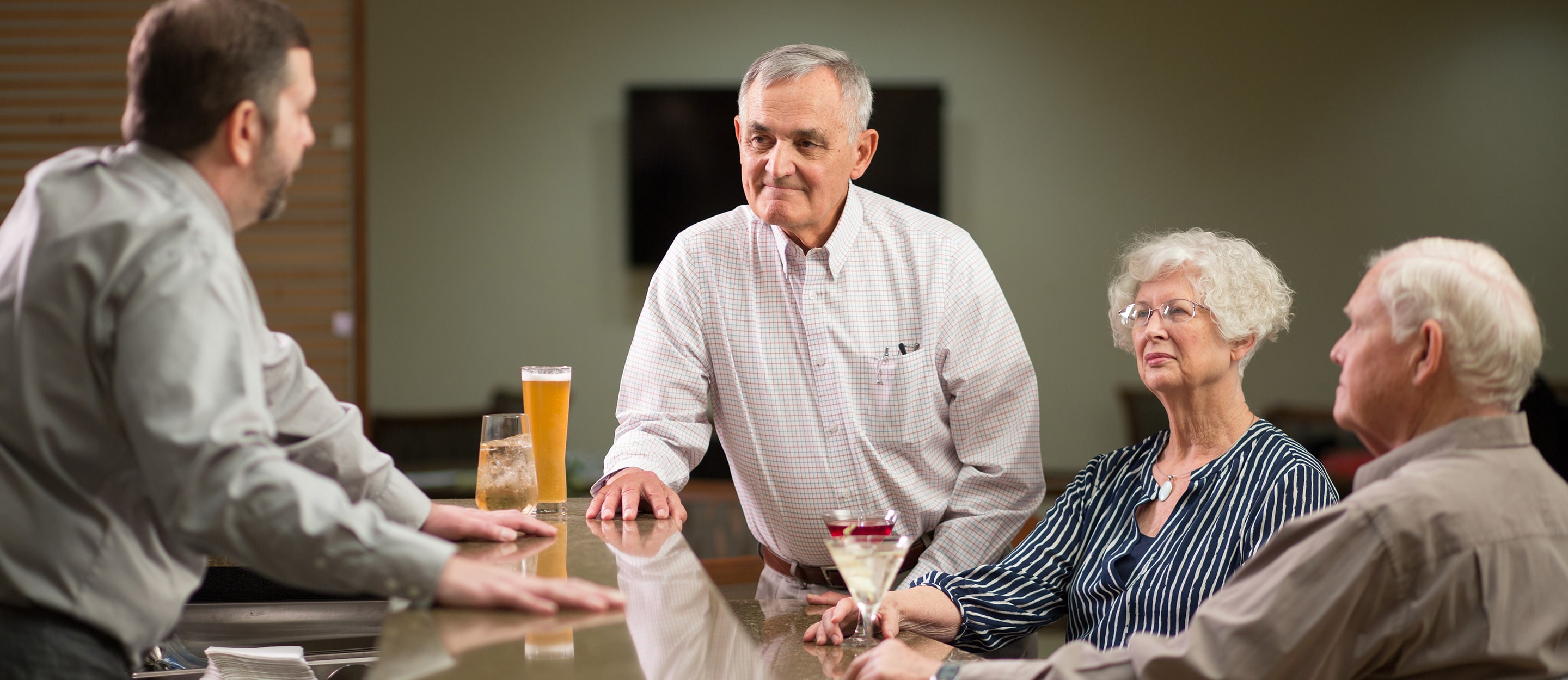 Three older adults sit at a bar facing a bartender. A man stands and listens attentively, while a woman and another man sit beside him with drinks, engaged in conversation in a relaxed, indoor setting.