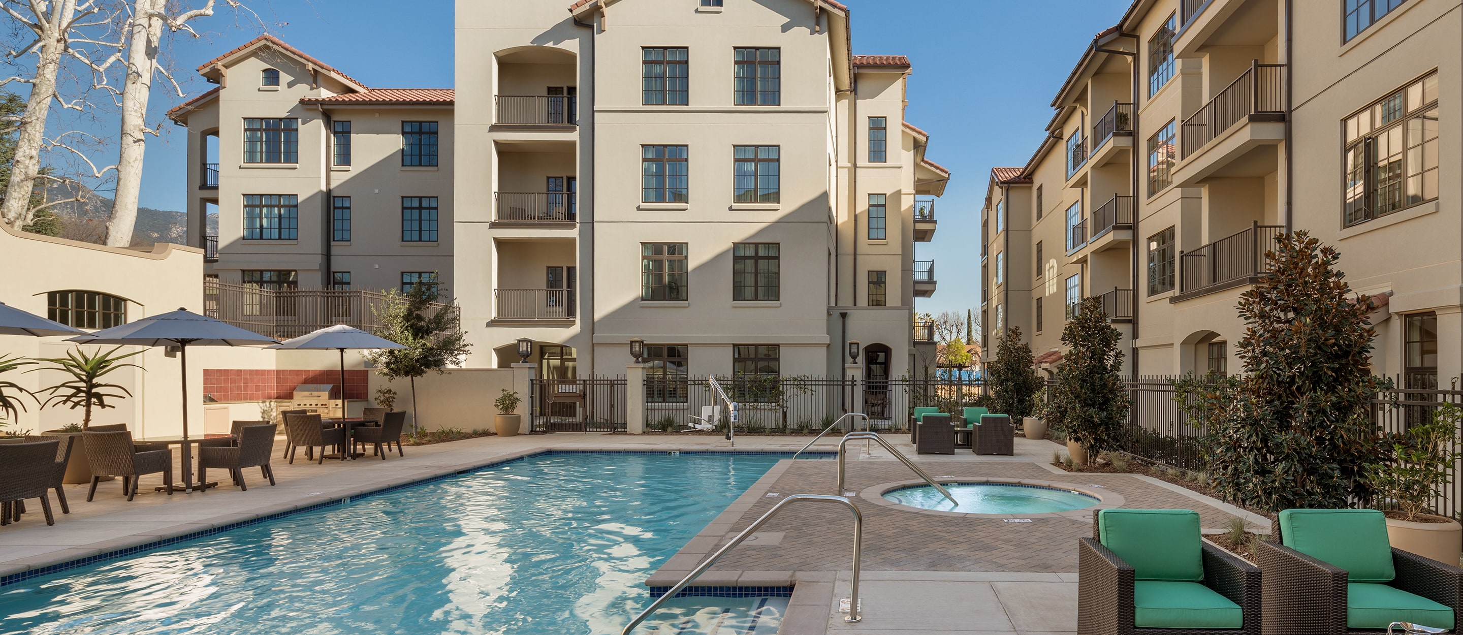 Modern apartment complex with beige buildings surrounding an outdoor swimming pool and hot tub. Poolside lounge chairs, tables with umbrellas, and green cushioned seats create a comfortable, inviting atmosphere under a clear sky.