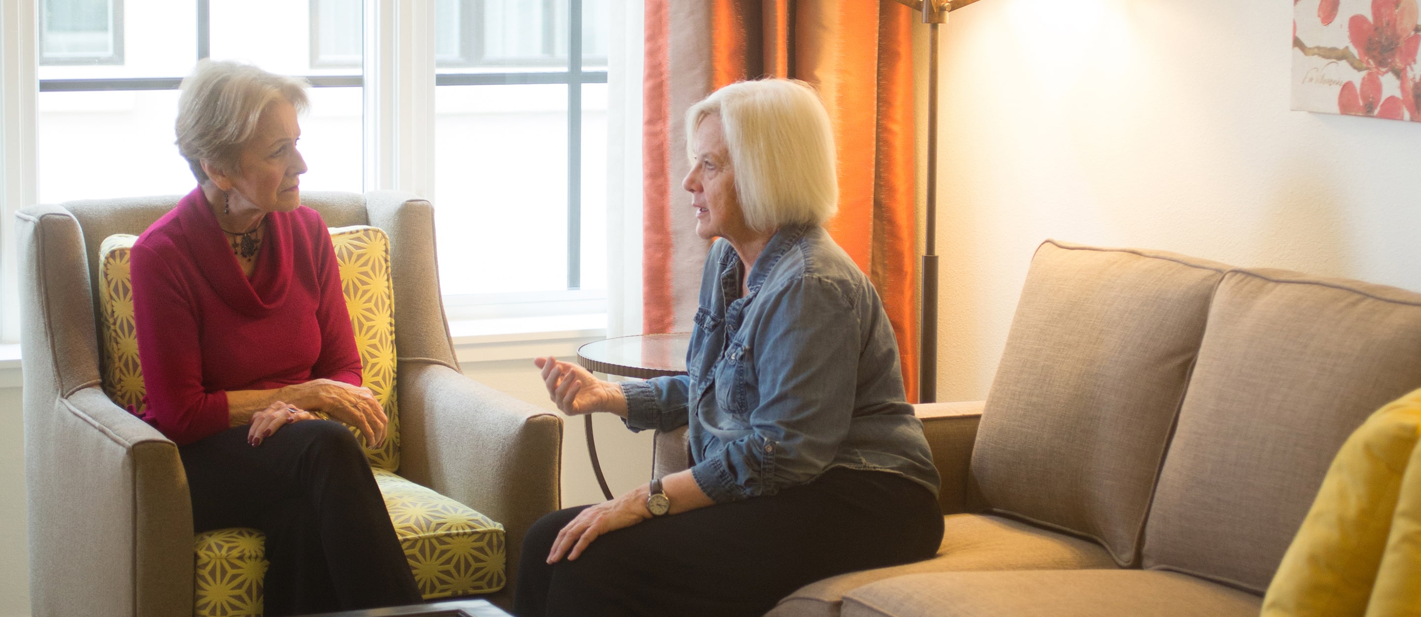Two older women sit in a well-lit living room having a serious conversation. One woman sits on an armchair, the other on a sofa, both facing each other and engaged in discussion.