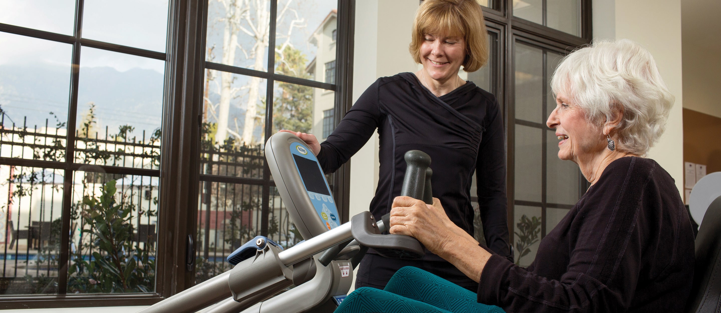 An older woman uses a seated exercise machine while a smiling woman stands beside her, offering guidance. They are indoors near large windows with a view of trees and buildings outside.
