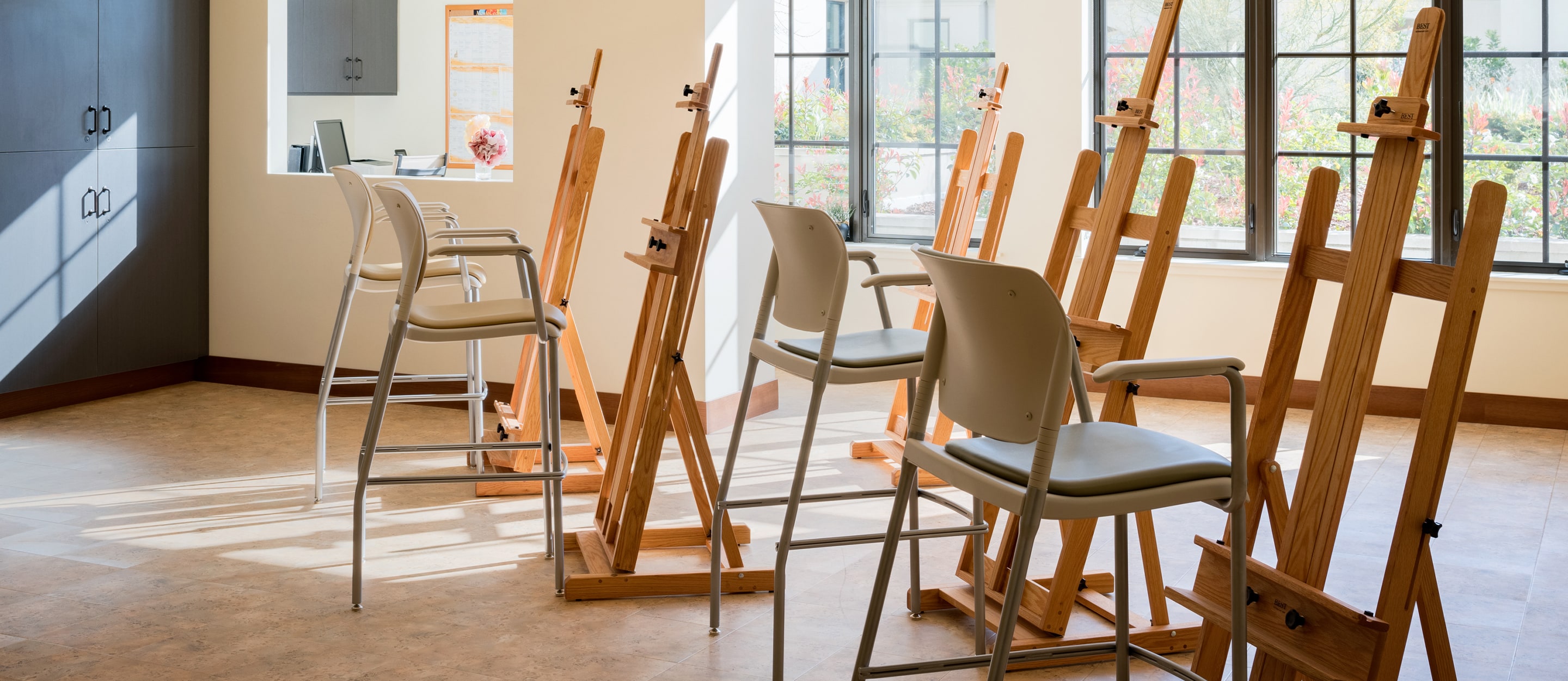 Several easels with chairs are arranged in a well-lit art studio with large windows, letting in natural light. The room has light-colored floors, cabinets, and a reception desk in the background.