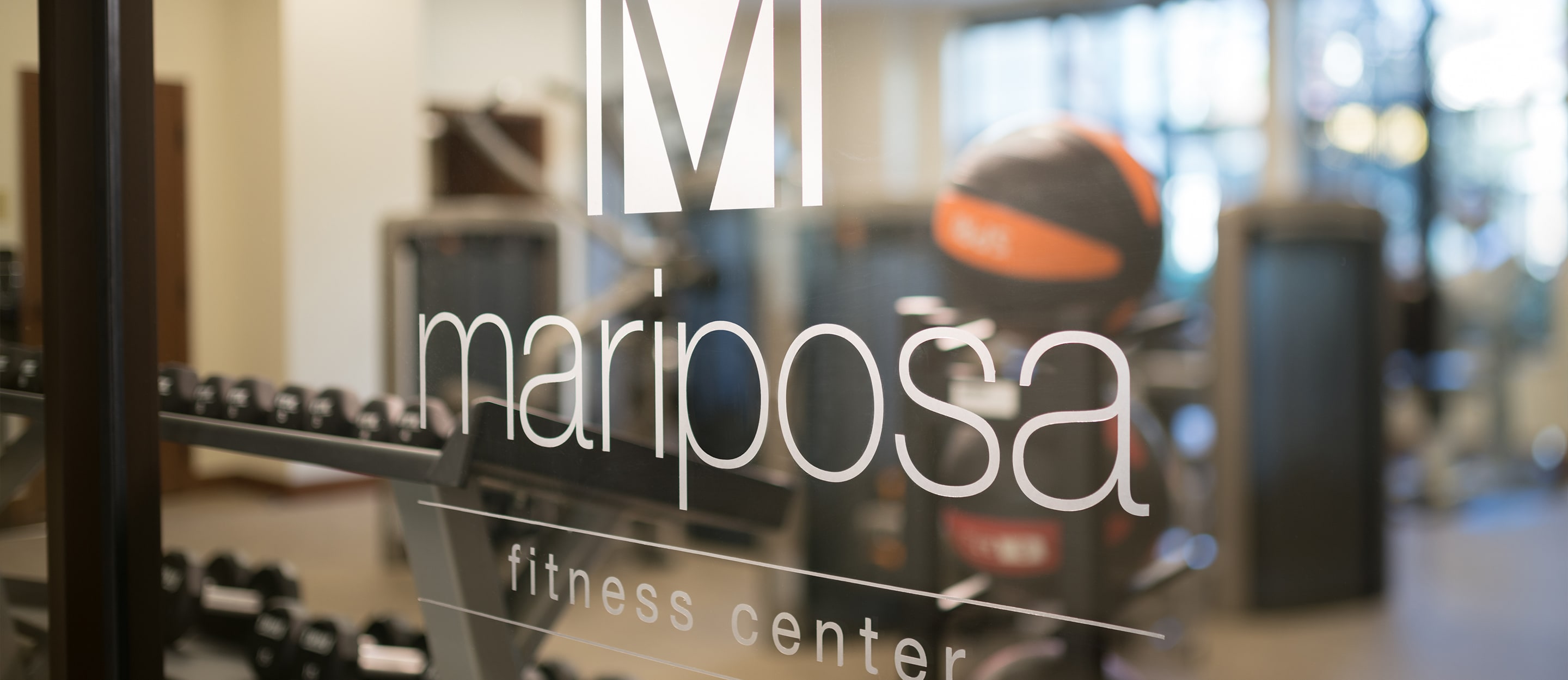 Glass door with mariposa fitness center and a large M logo, with a blurred view of gym equipment like dumbbells and medicine balls visible in the background.