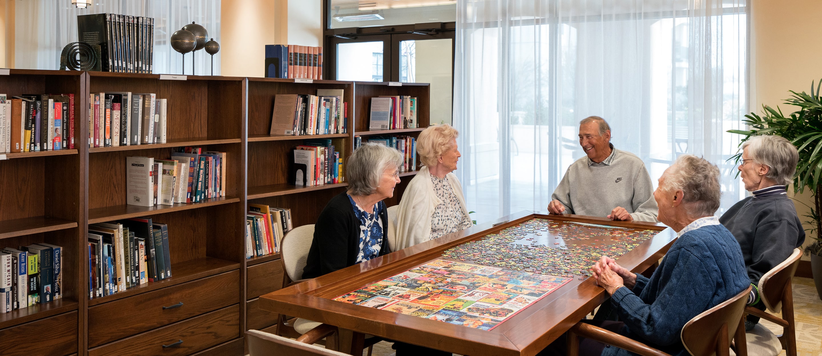 Five older adults sit around a table working on a colorful jigsaw puzzle in a bright room with bookshelves and large windows in the background.