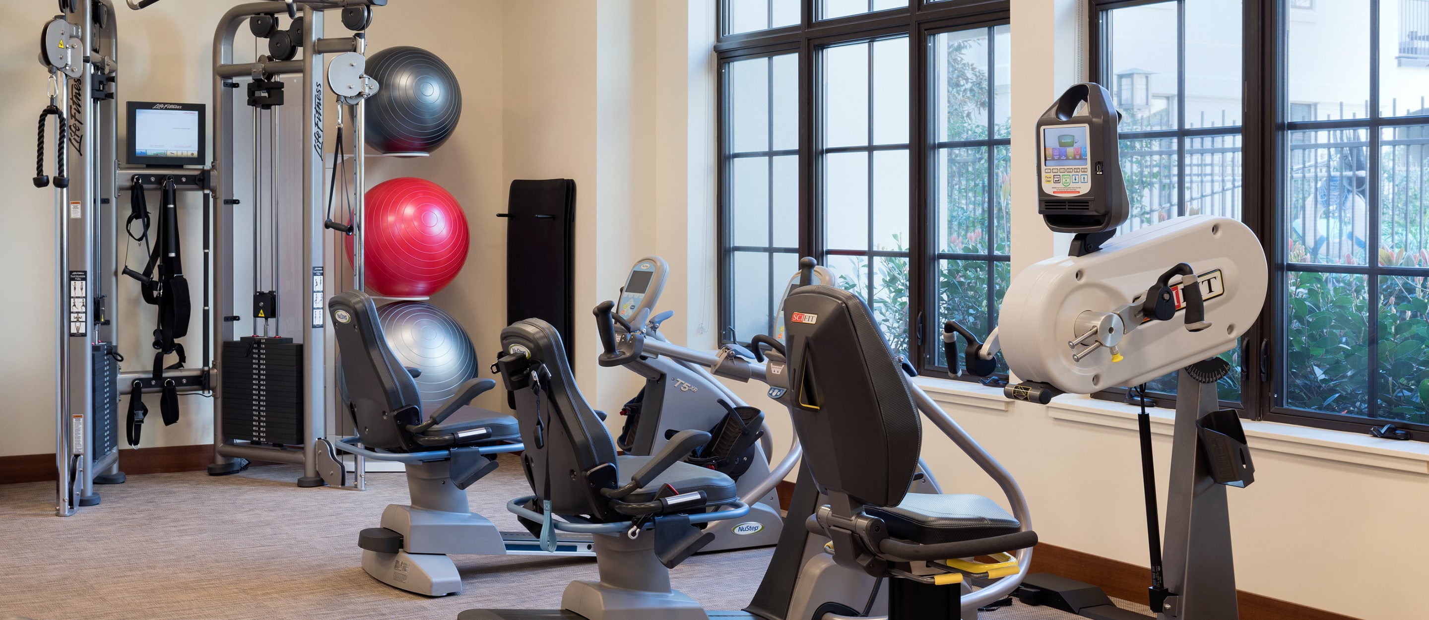 A modern gym with exercise machines, including recumbent bikes, a cable machine, and a hand cycle. Large windows let in natural light. Colorful stability balls are stacked in a corner.