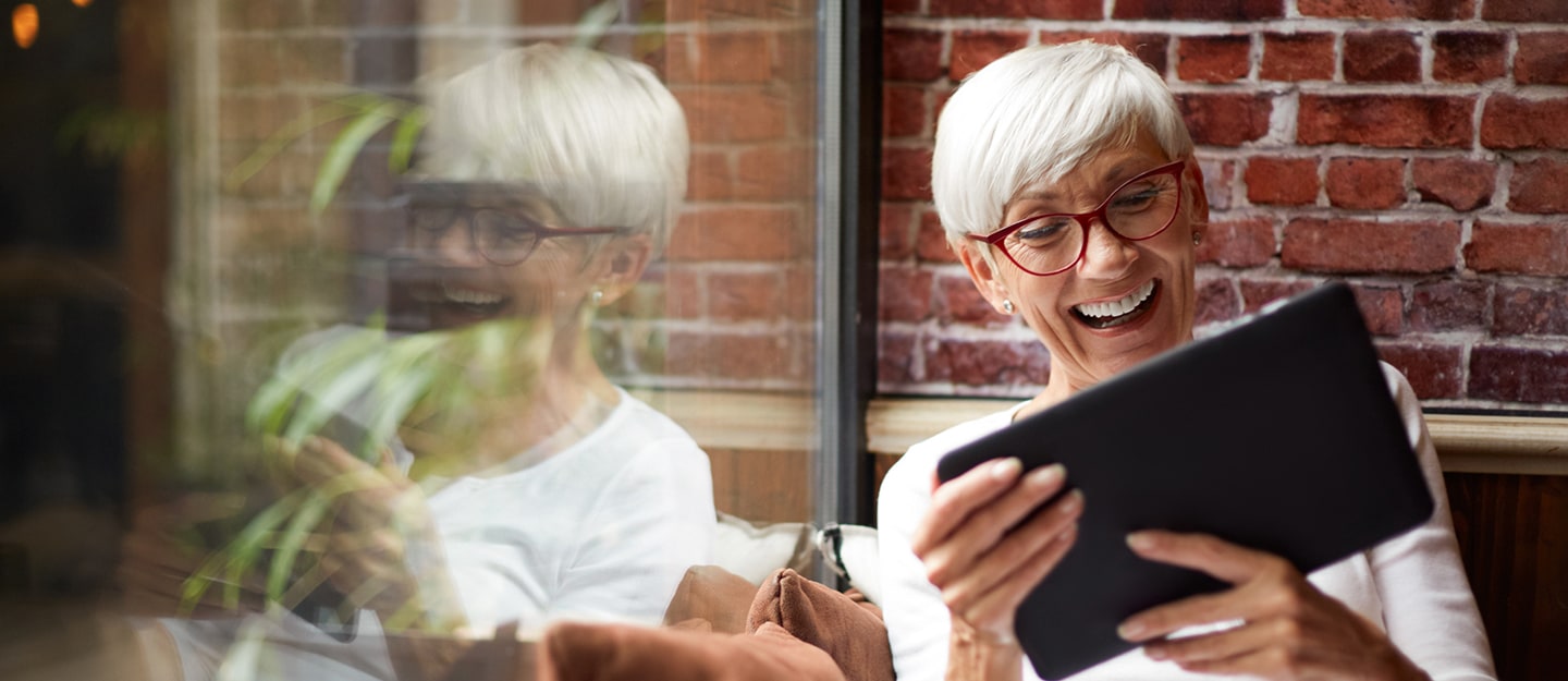 An older woman with short white hair and red glasses smiles while using a tablet, sitting by a window with a brick wall in the background. Her reflection is visible in the glass.