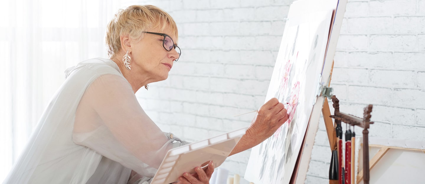 An older woman with short blonde hair and glasses is painting on a canvas in a bright room, holding a palette and brush. She is focused on her artwork, surrounded by painting supplies.