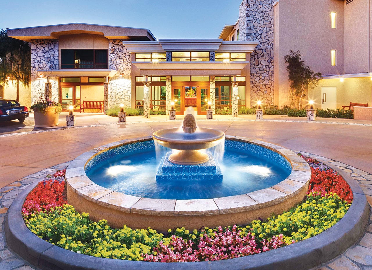 A circular fountain surrounded by colorful flowers sits in front of a modern building with large windows and stone walls, illuminated in the evening.