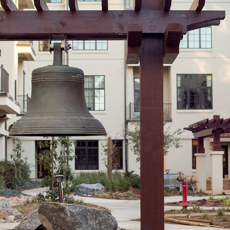 A large bronze bell hangs from a wooden pergola structure in a courtyard with beige buildings, windows, greenery, and stone landscaping in the background.