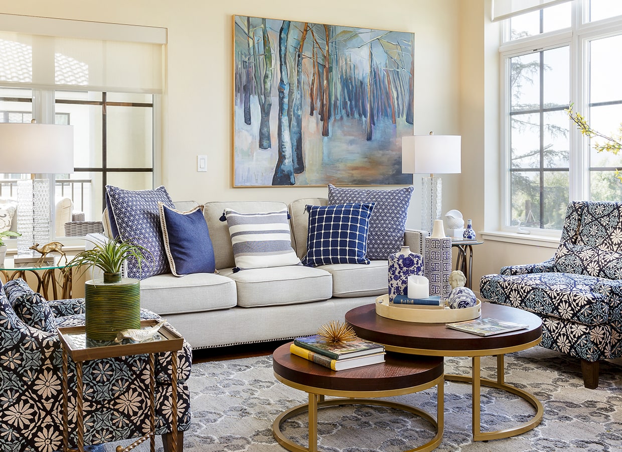 Bright living room with a beige sofa, blue and white patterned pillows, two matching armchairs, round coffee tables with books and decor, a potted plant, and a large abstract forest painting on the wall.