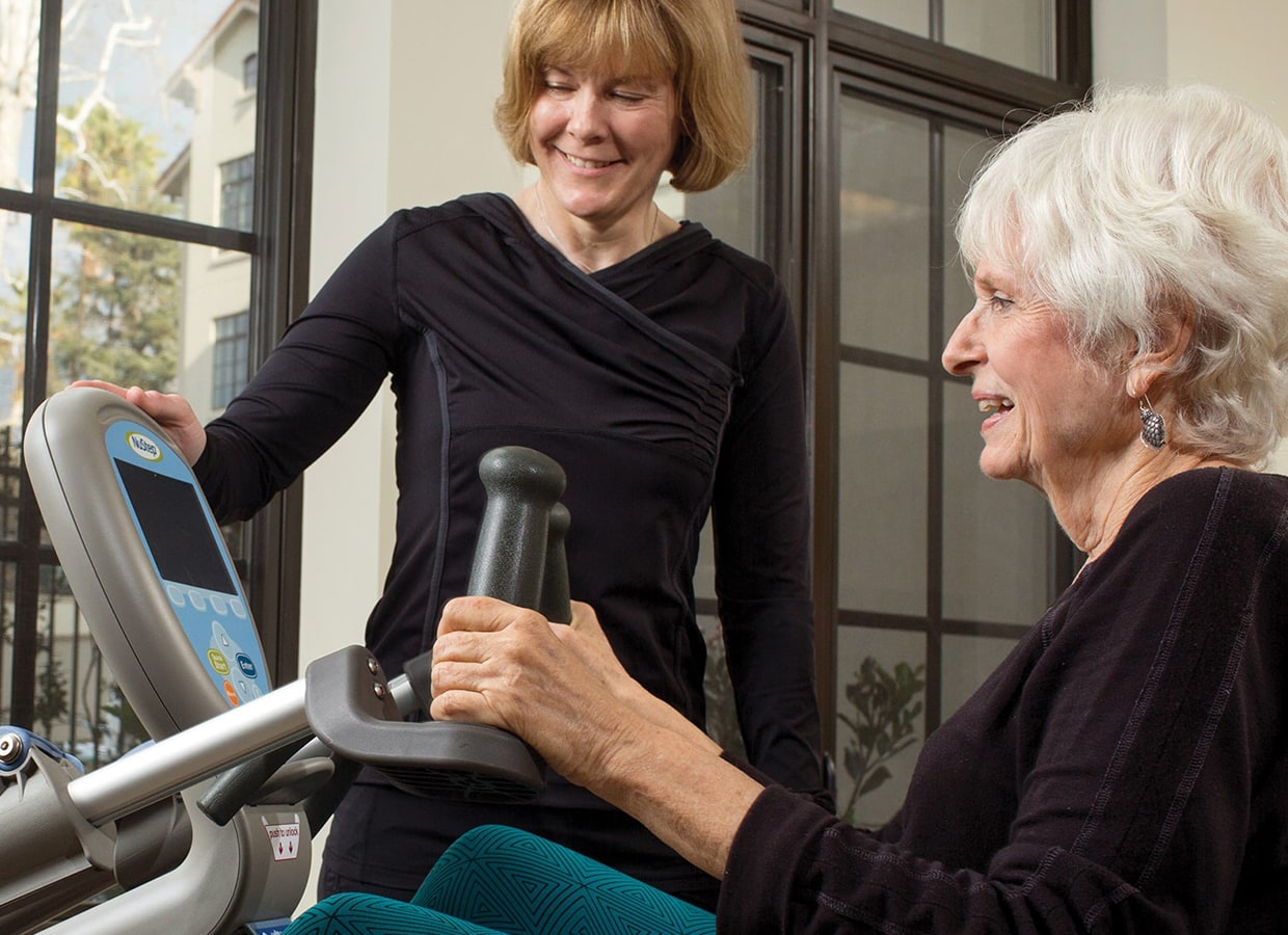 An older woman exercises on a stationary machine while a younger woman beside her smiles and provides guidance in a bright room with large windows.