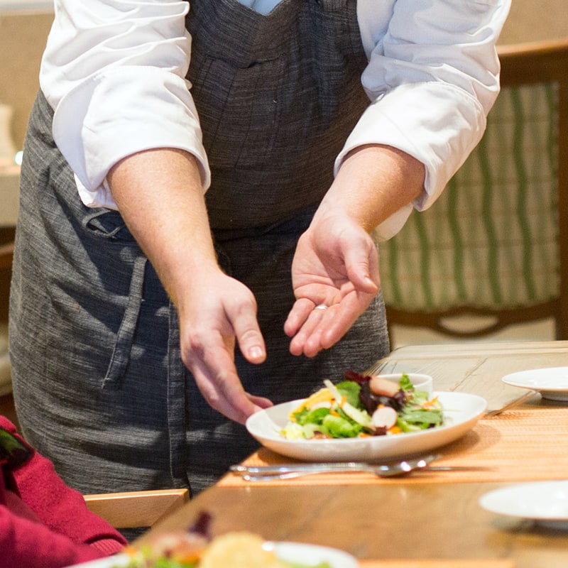 A person in a white shirt and gray apron is serving a plate of salad on a table set for a meal. Only their torso and arms are visible. The table has a placemat and other dishes.