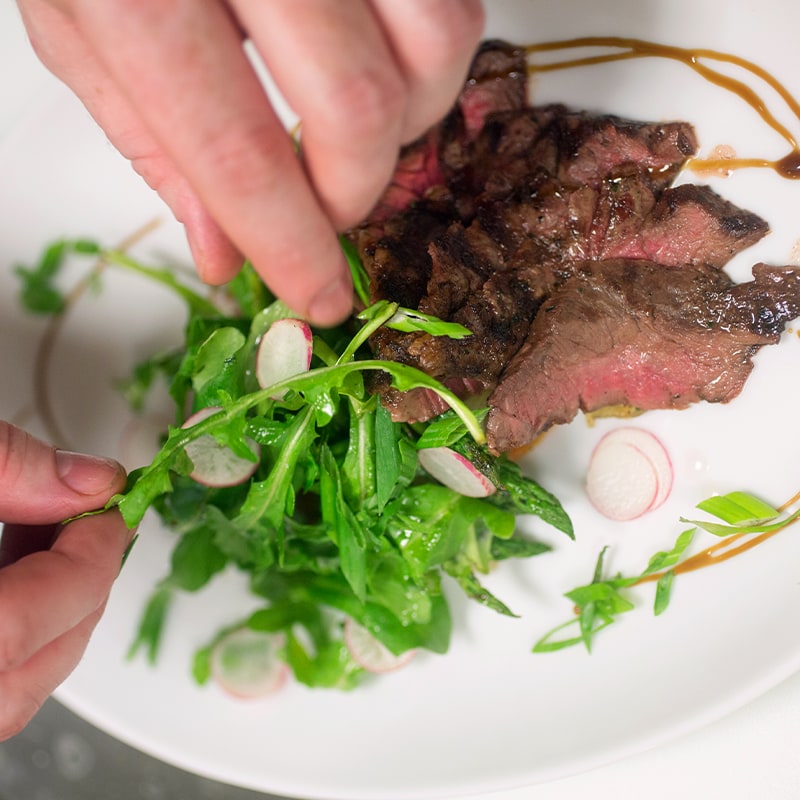 A close-up of hands plating a dish with sliced medium-rare steak, arugula salad, and thinly sliced radishes on a white plate, garnished with a drizzle of sauce.