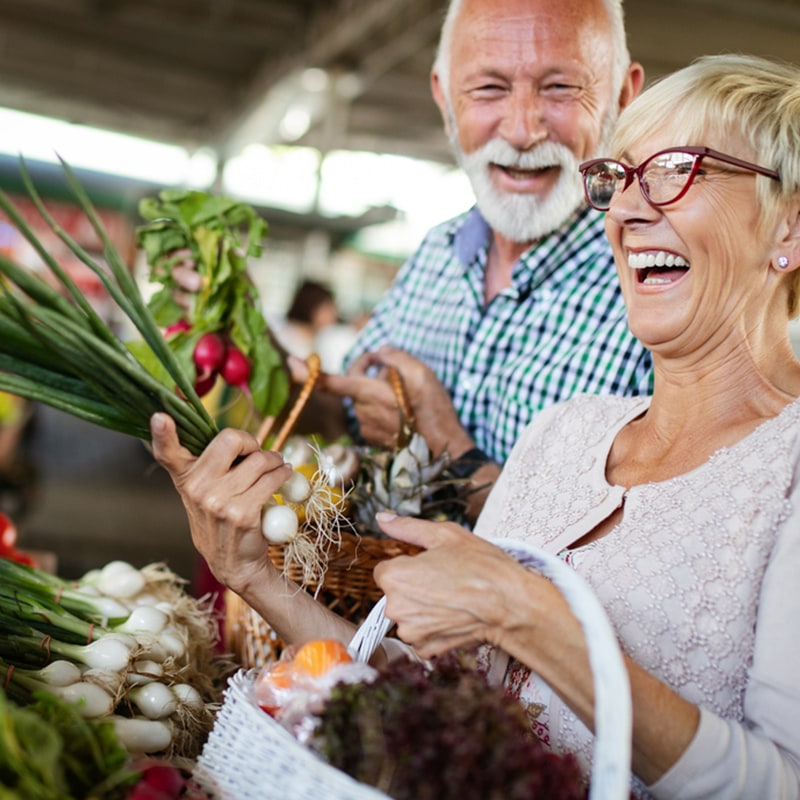 An older couple smiles and laughs while shopping for fresh vegetables at a market, holding baskets filled with produce such as onions, radishes, and greens.