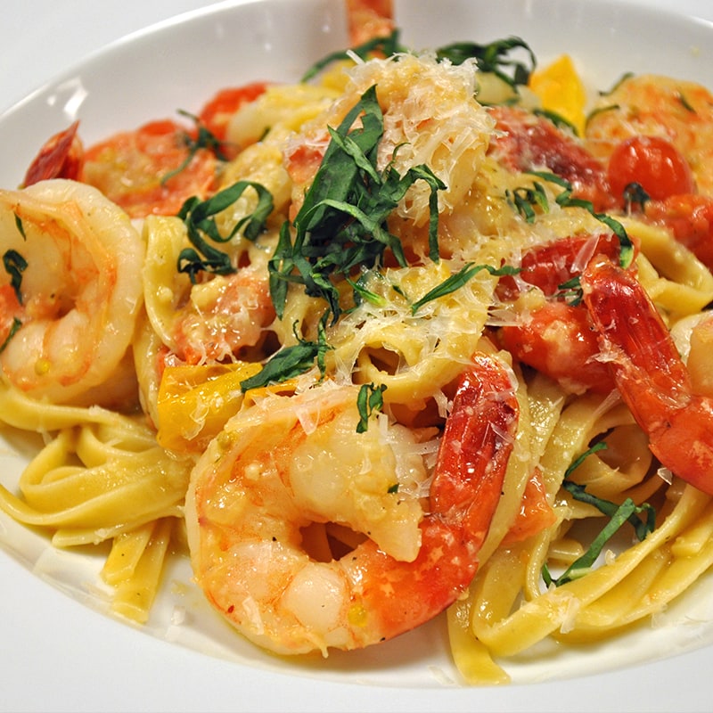 A close-up of shrimp pasta topped with grated cheese and fresh herbs, featuring large shrimp, fettuccine noodles, and pieces of yellow bell pepper in a creamy tomato sauce on a white plate.