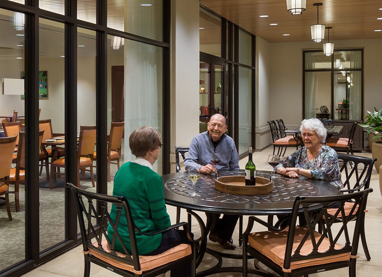 Three elderly people sit around a round metal table on a covered patio, talking and smiling. A wine bottle and glasses are on the table. The patio is next to a large window showing a dining area inside.
