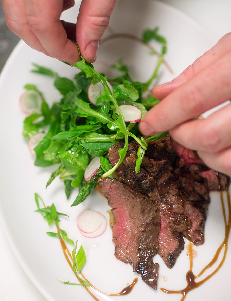 With creativity, a person arranges fresh arugula and radish slices on a plate next to sliced grilled steak, garnished with a drizzle of sauce.