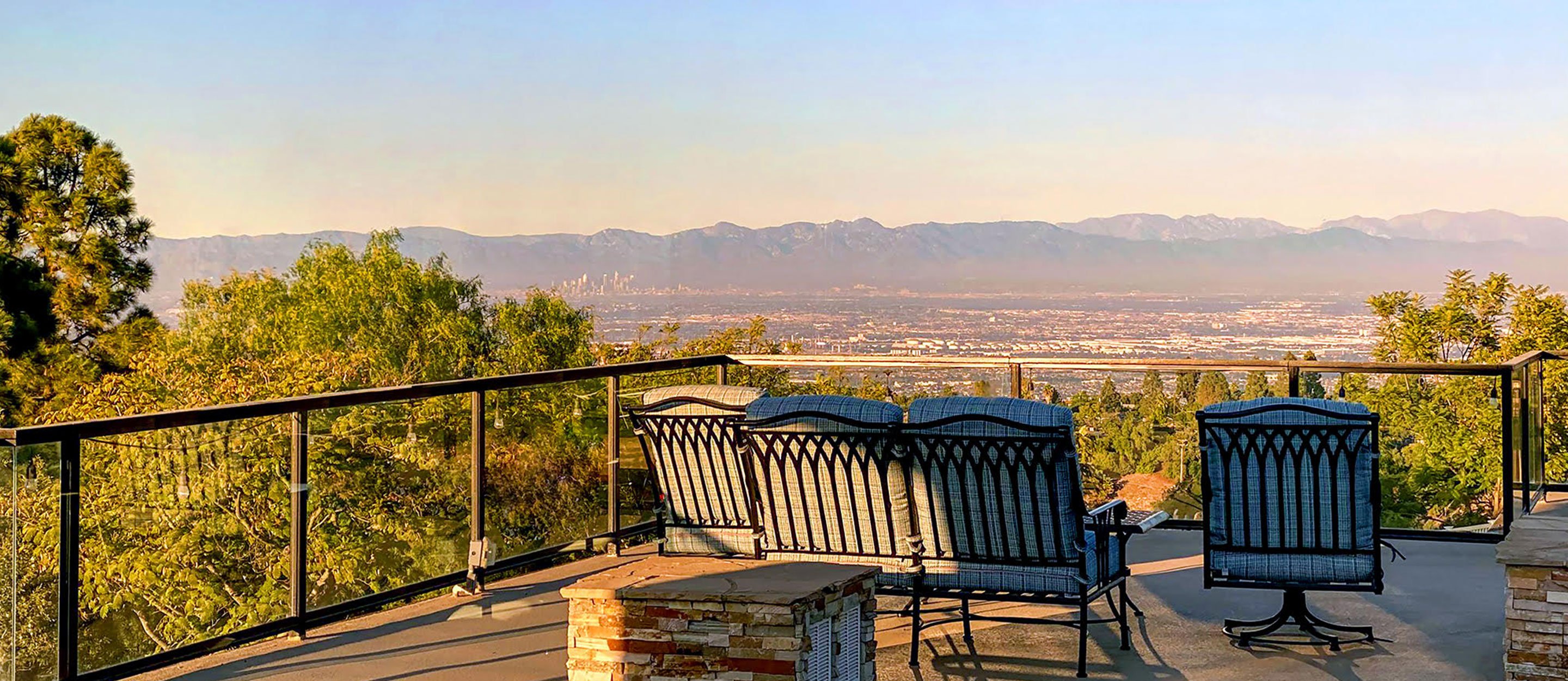 A patio with metal chairs and a glass railing overlooks a scenic valley and distant mountains under a clear sky, surrounded by trees and greenery.