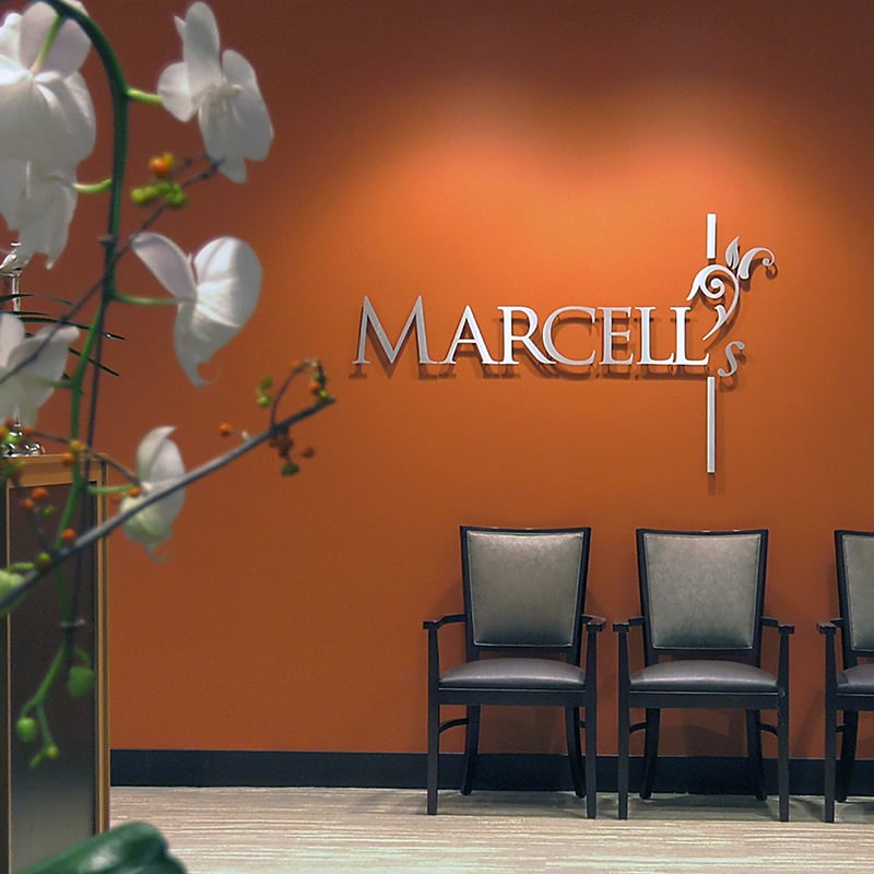 Three empty chairs line up against an orange wall with the silver sign Marcells. White orchids are in the foreground on the left side of the image. The setting appears to be a waiting area or lobby.