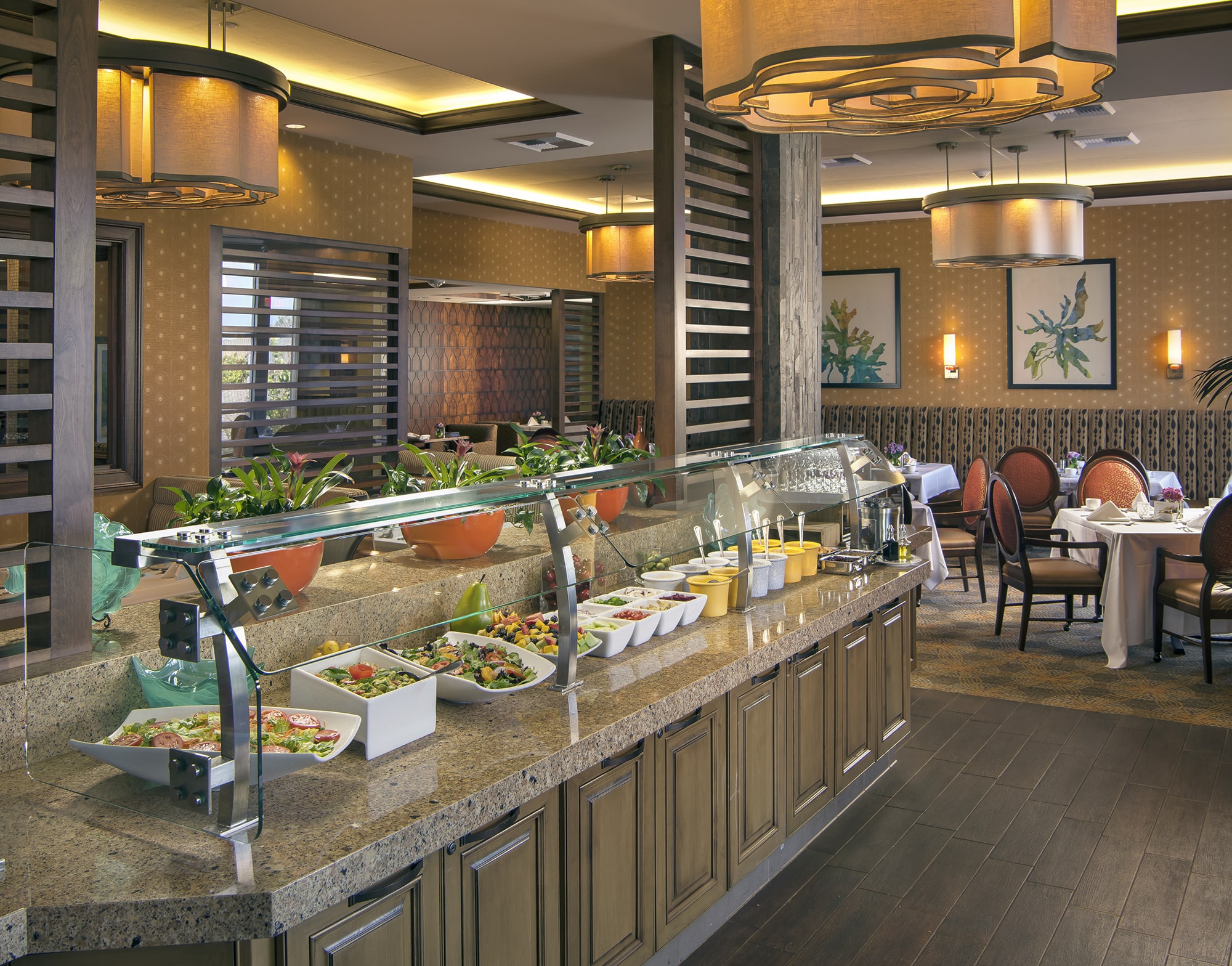 A buffet with various salads and toppings sits on a granite counter in an elegant restaurant with wooden floors, decorative lighting, and tables set for dining in the background.