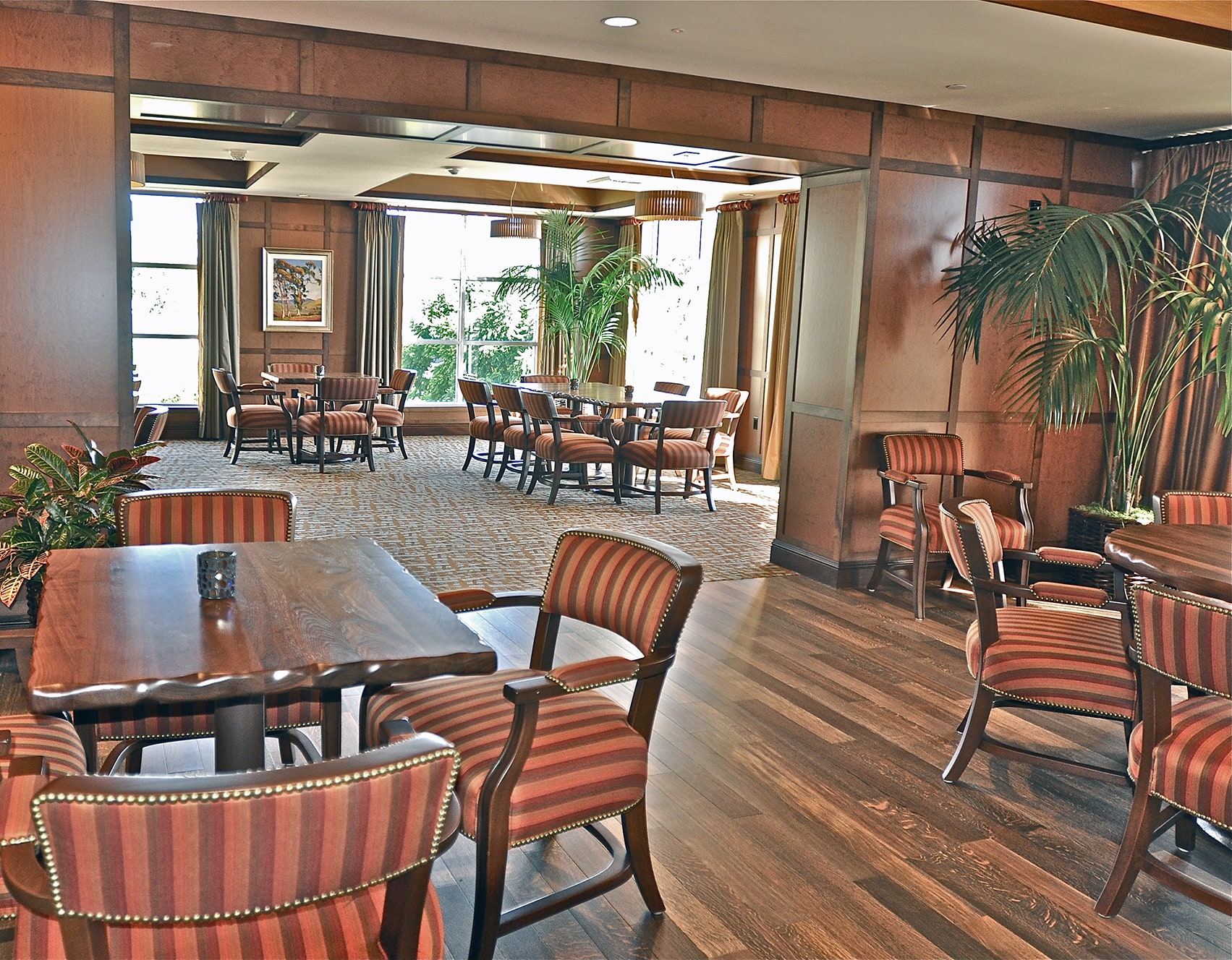 A spacious dining room with wooden floors, wood-paneled walls, and multiple tables with striped upholstered chairs. Large windows and potted plants create a bright, inviting atmosphere.