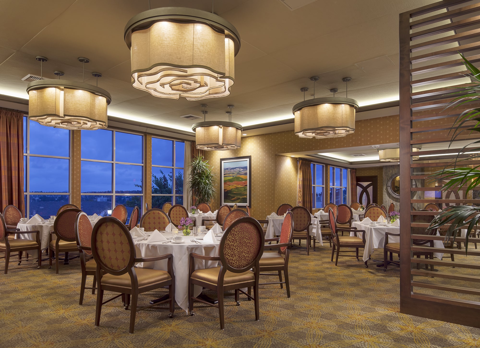 Elegant restaurant dining room with round tables covered in white tablecloths, cushioned chairs, large decorative ceiling lights, and floor-to-ceiling windows overlooking a scenic view at dusk. A painting hangs on the wall.