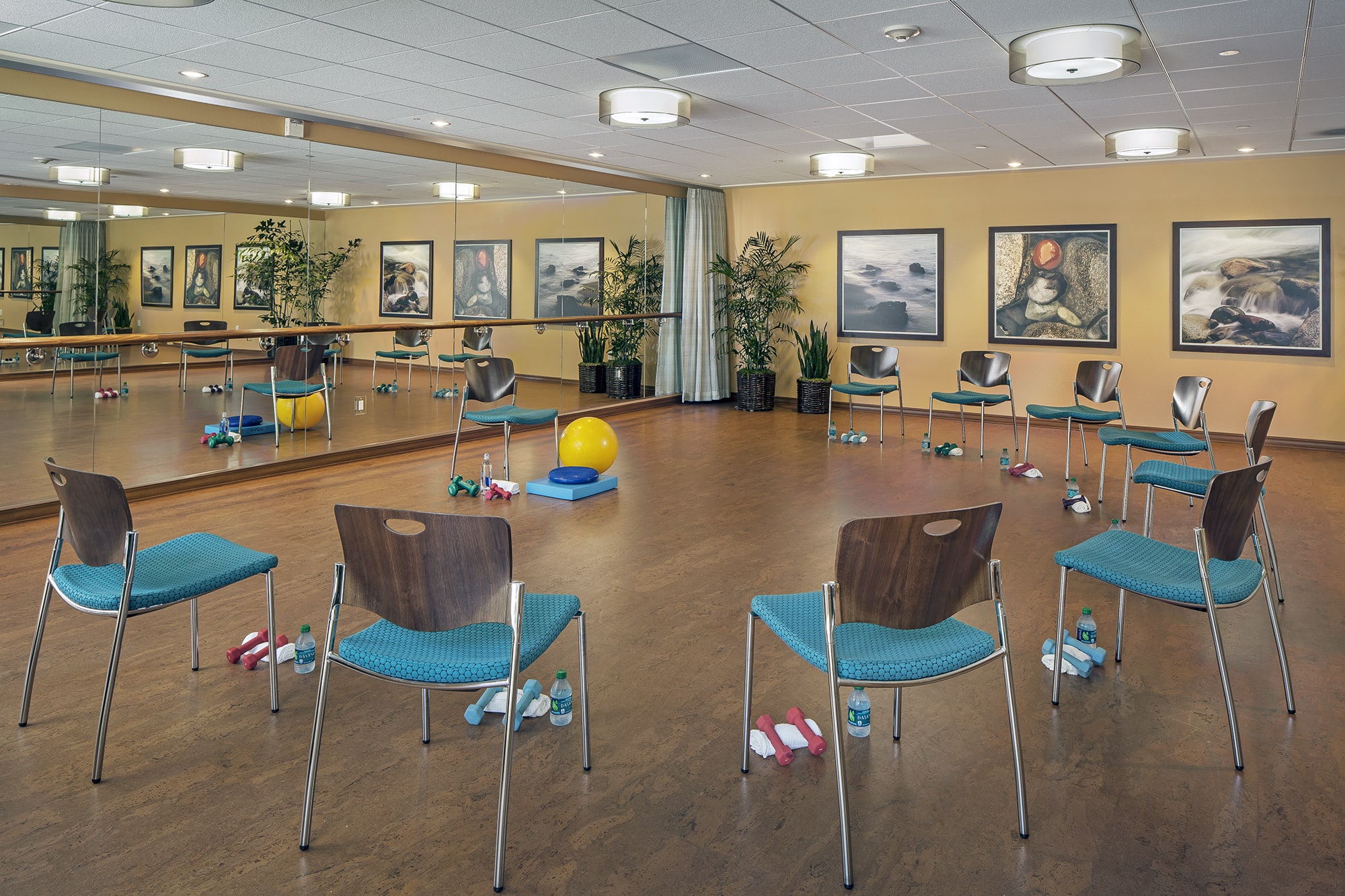 A circle of chairs with blue seats is arranged in a room with wooden floors. Each chair has water bottles, towels, and small dumbbells nearby. There are mirrors, large plants, and framed artwork on the walls.
