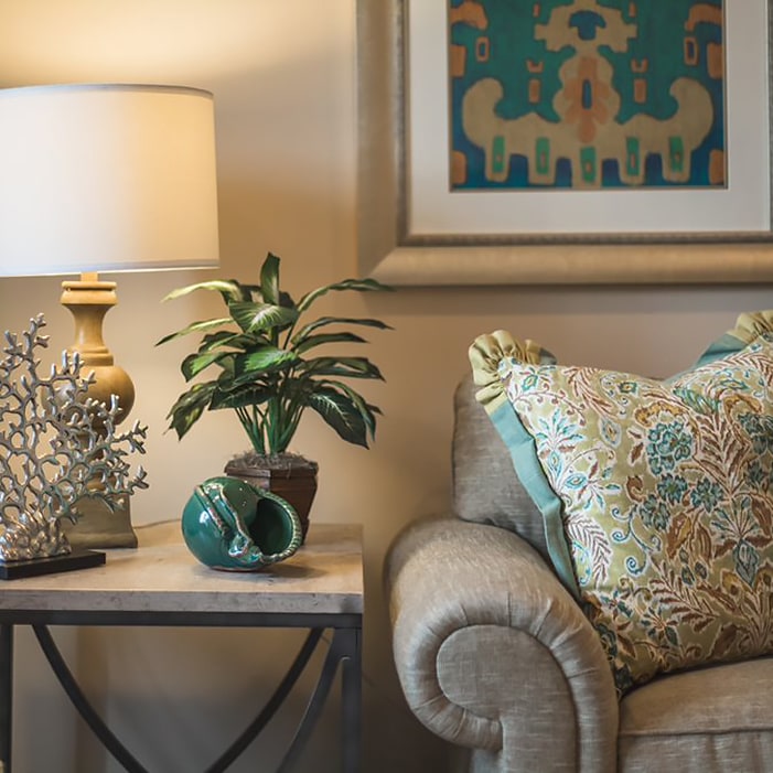 A cozy living room corner with a beige sofa adorned with patterned pillows, a side table holding a gold lamp, a small potted plant, and decorative items, with a colorful framed artwork on the wall.