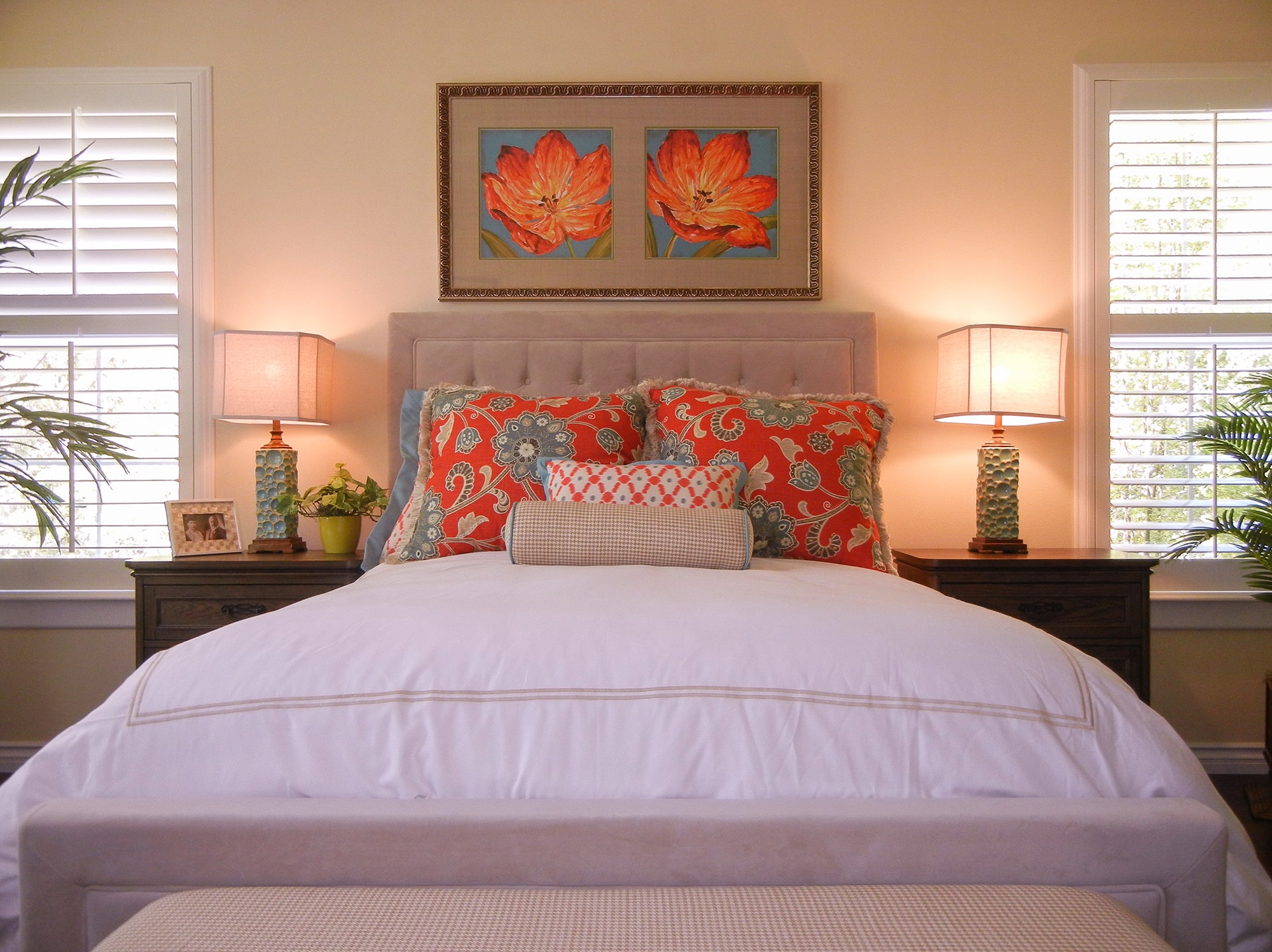 A neatly made bed with colorful orange and gray floral pillows, a white comforter, and a decorative bolster pillow. There are two lamps on wooden nightstands, plants, and a framed orange flower artwork above the headboard.