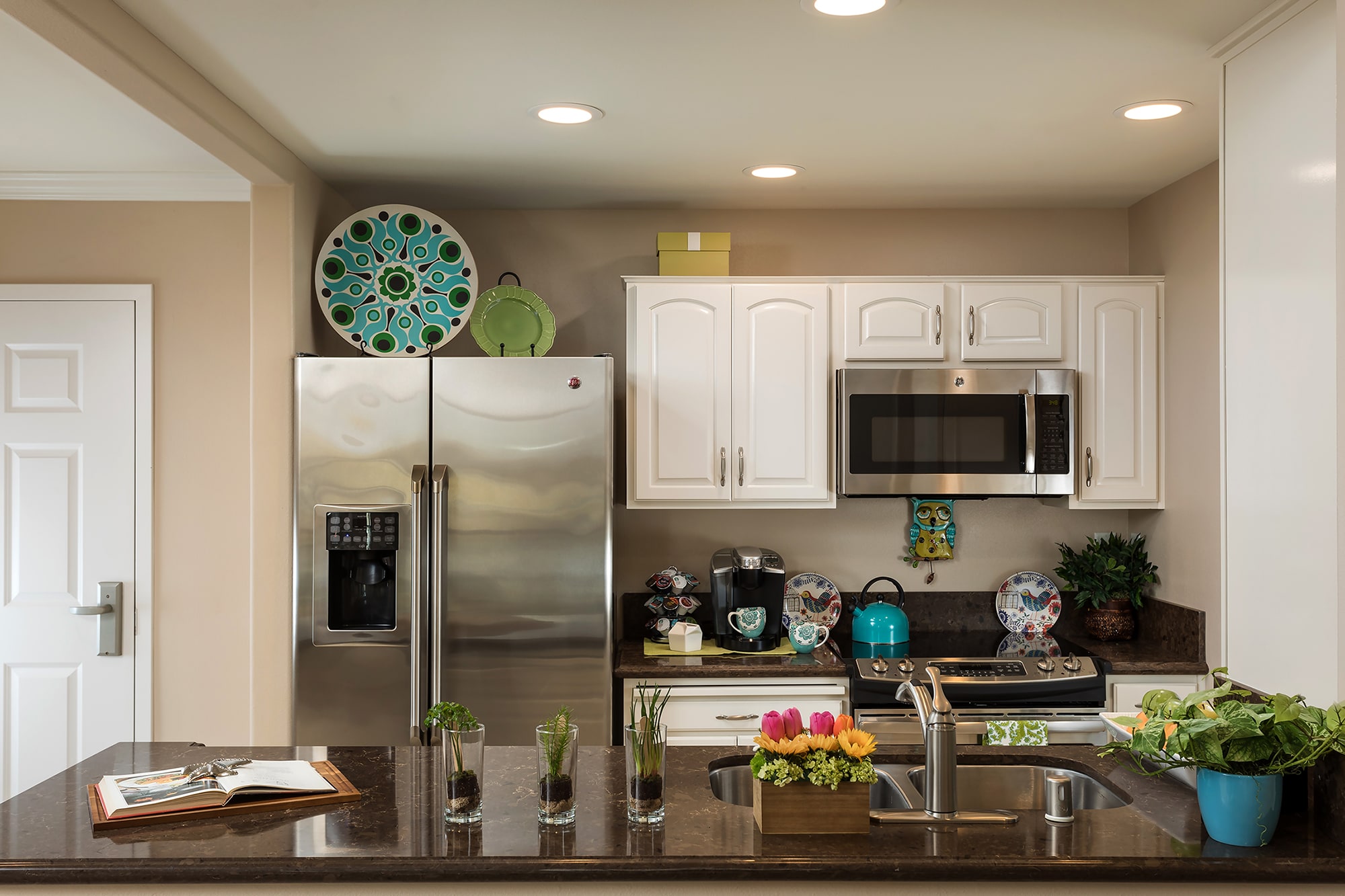 A modern kitchen with white cabinets, stainless steel appliances, decorative plates, potted plants, and a brown countertop with an open cookbook and a small flower arrangement.