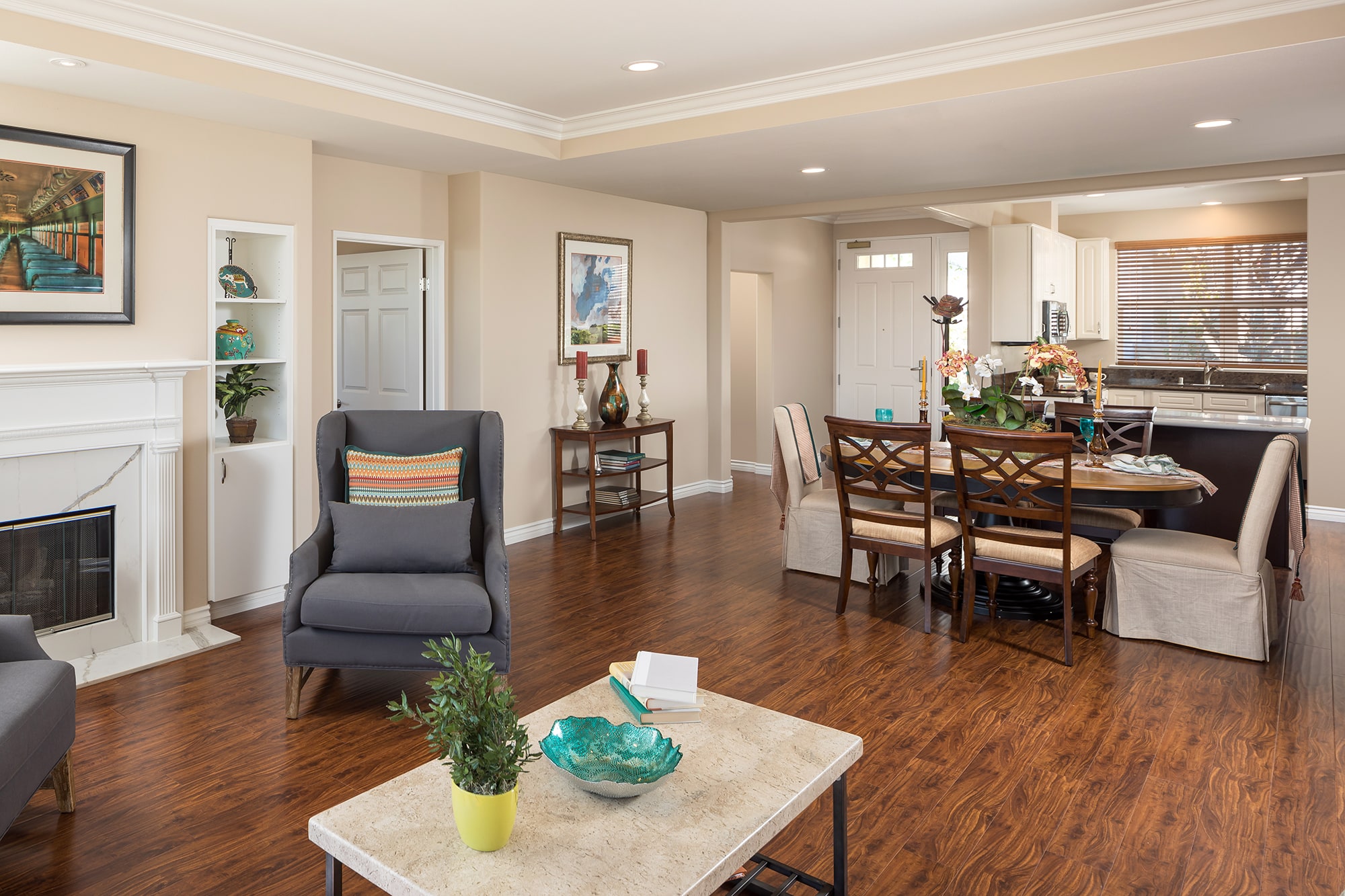 Open-concept living and dining area with wood floors, modern furniture, a fireplace, and kitchen in the background. Light walls and decor give a bright, welcoming atmosphere.