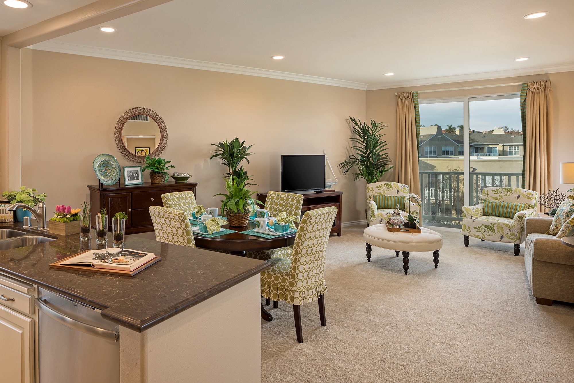 Open-concept living and dining area with beige walls, cozy seating, green accents, plants, a dining table set for four, TV, and large window with curtains letting in natural light.