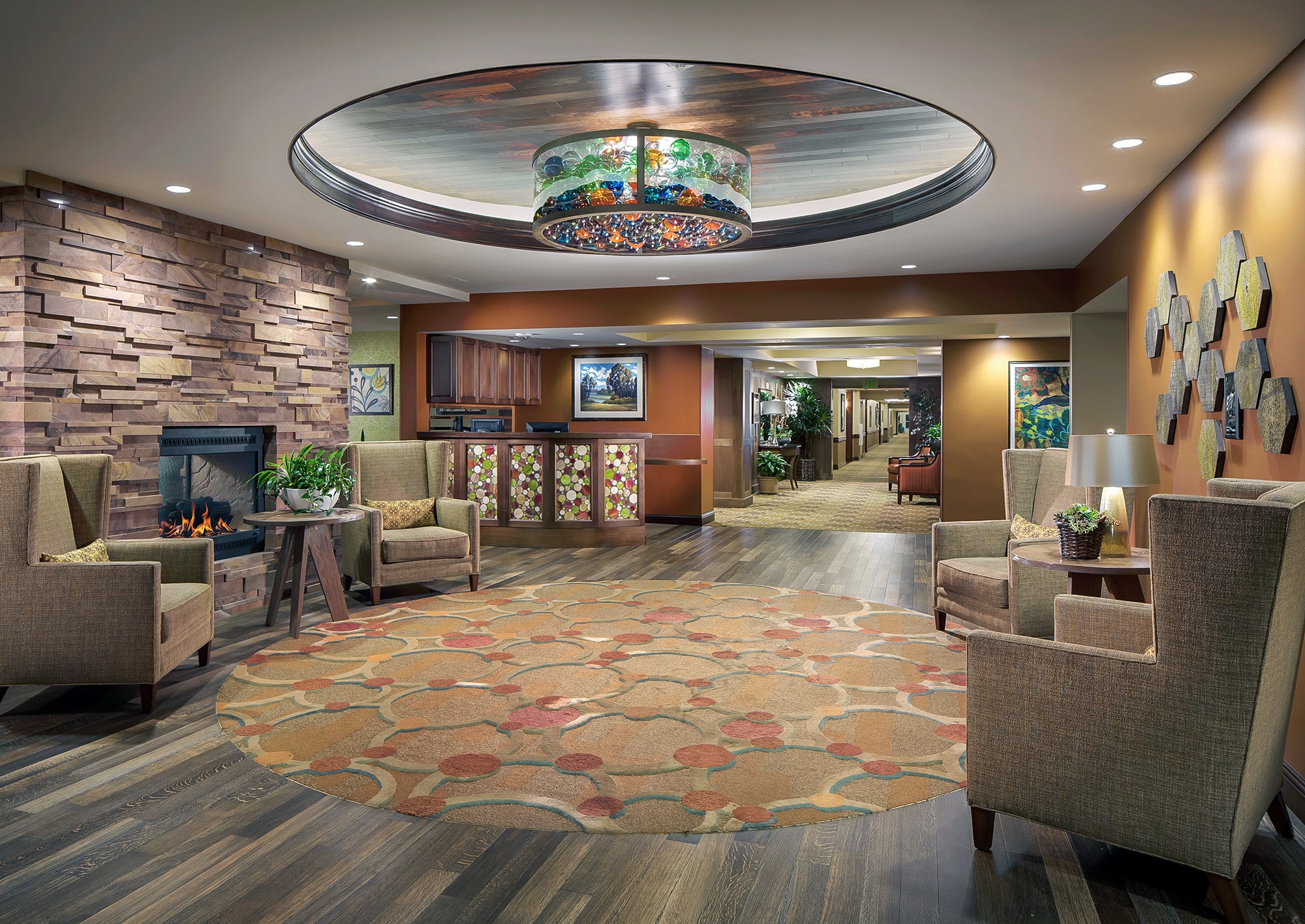 A modern, cozy lobby with armchairs, a stone fireplace, colorful art, a round decorative rug, and a circular ceiling light fixture. There are wood floors, soft lighting, and plants in the background hallway.