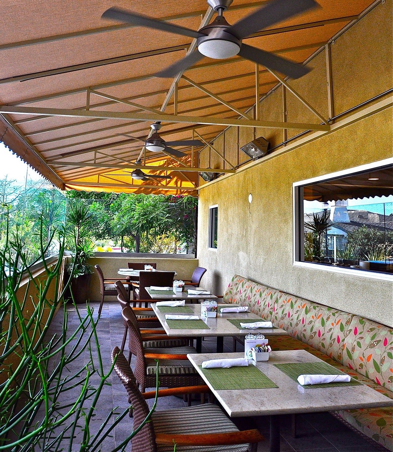 Outdoor restaurant patio with ceiling fans, covered by a tan awning. Tables are set with napkins and placemats. Cushioned benches with floral patterns line one side; greenery and trees are visible outside.