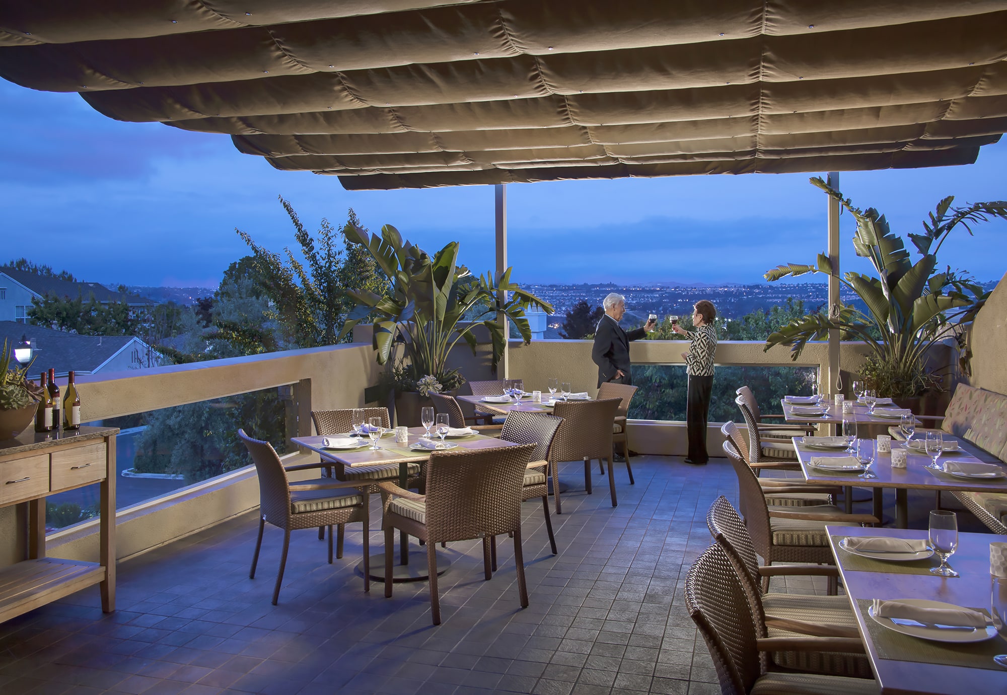 Outdoor restaurant patio at dusk with set tables, potted plants, and a scenic city view. Two people stand by the railing, conversing and enjoying the view. Warm lighting creates a cozy, inviting atmosphere.
