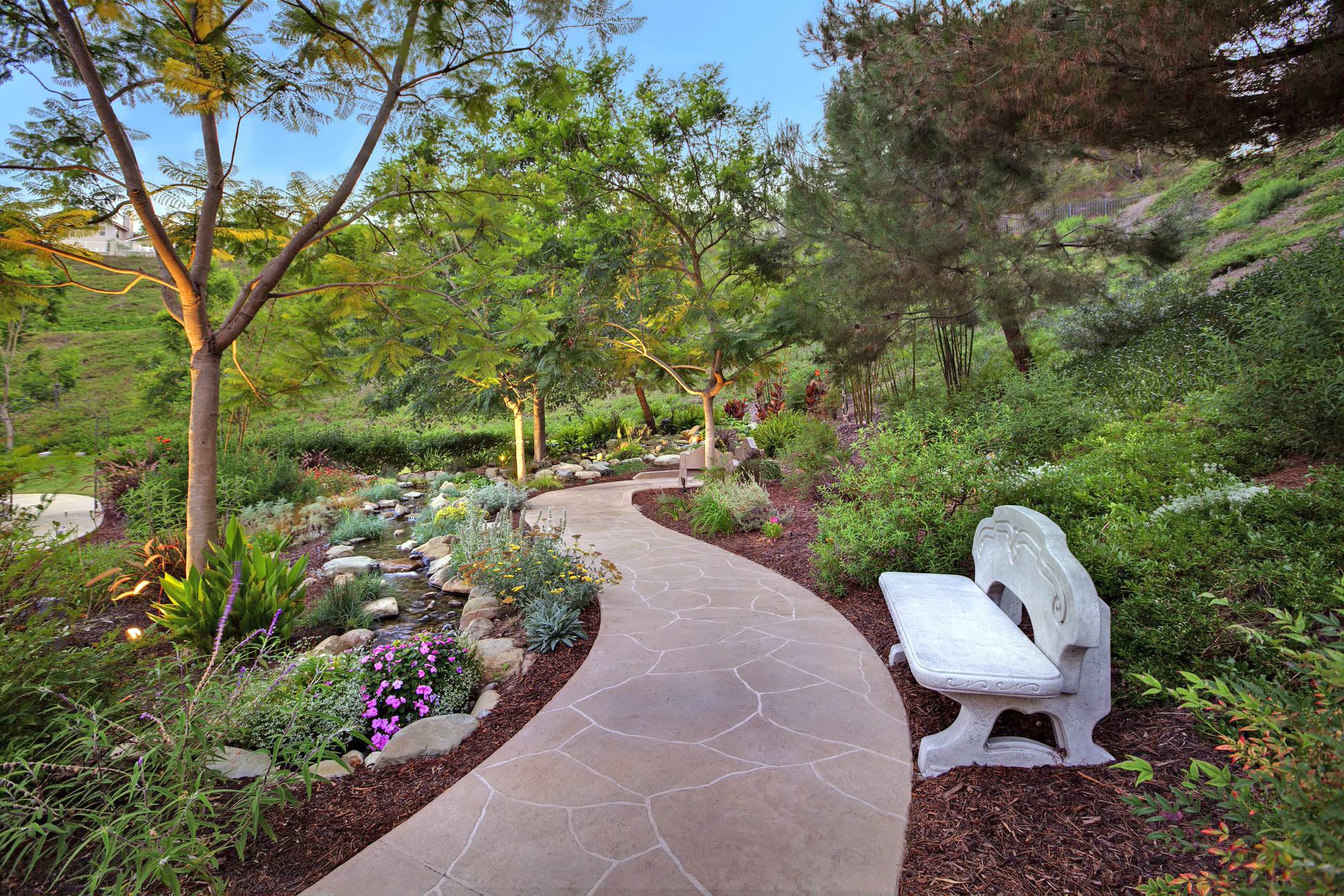 A stone path curves through a lush garden with green trees, colorful flowers, and shrubs. A white stone bench sits beside the path, inviting visitors to rest and enjoy the peaceful, natural setting.