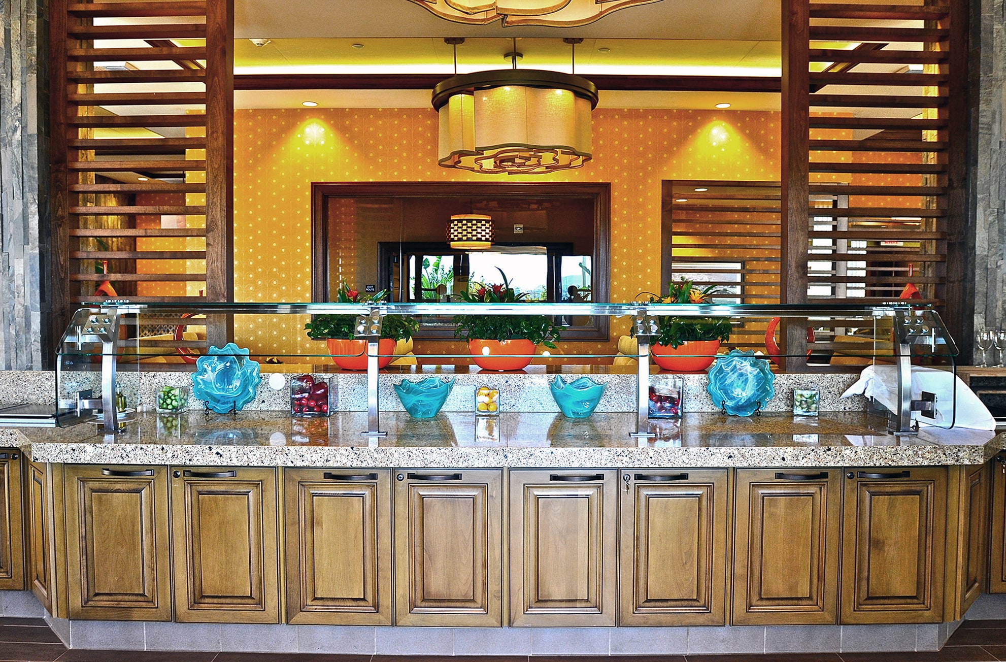 A buffet counter with wooden cabinets, glass sneeze guard, colorful bowls filled with salad ingredients, and decorative blue glass sculptures, set in a well-lit, elegant dining area.