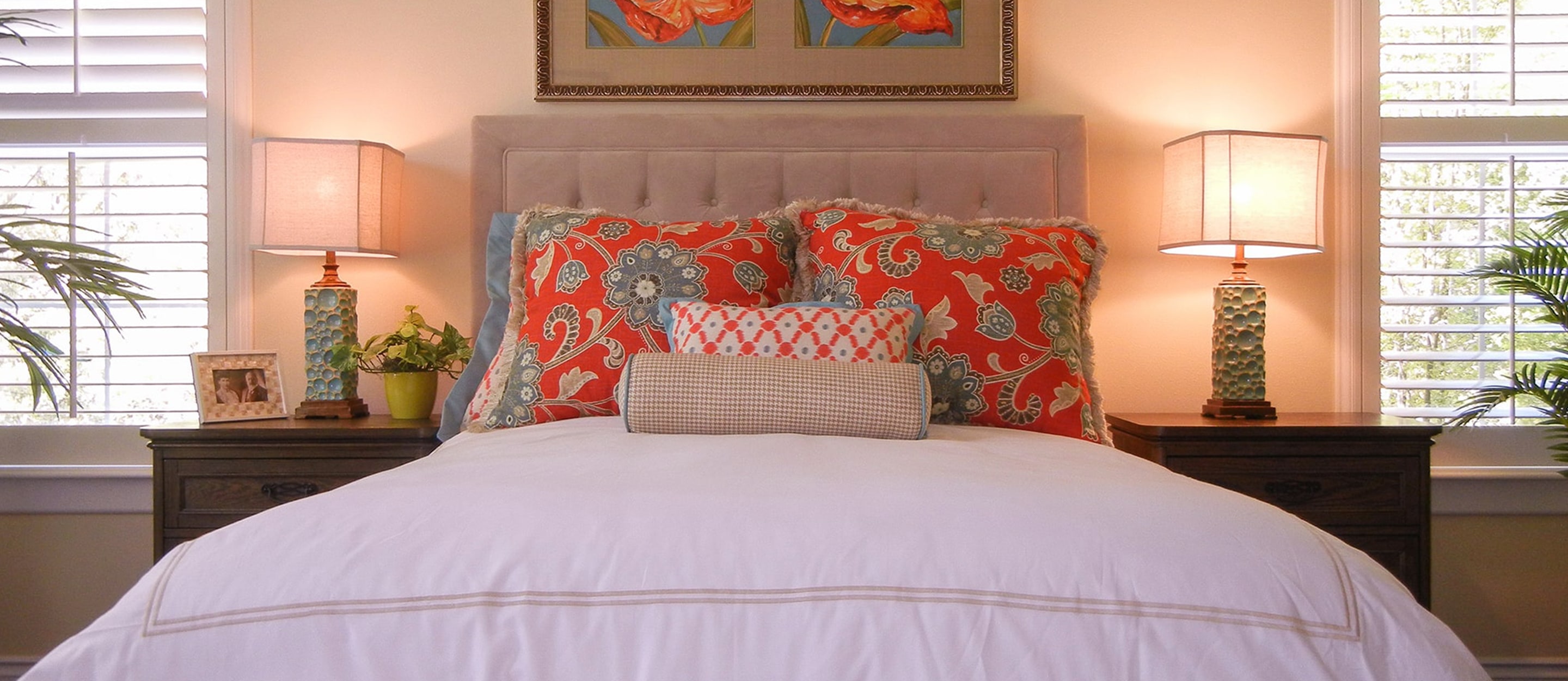A cozy bedroom with a neatly made bed, white comforter, colorful patterned pillows, matching bedside tables, two lamps, shuttered windows, and a framed floral painting above the headboard.