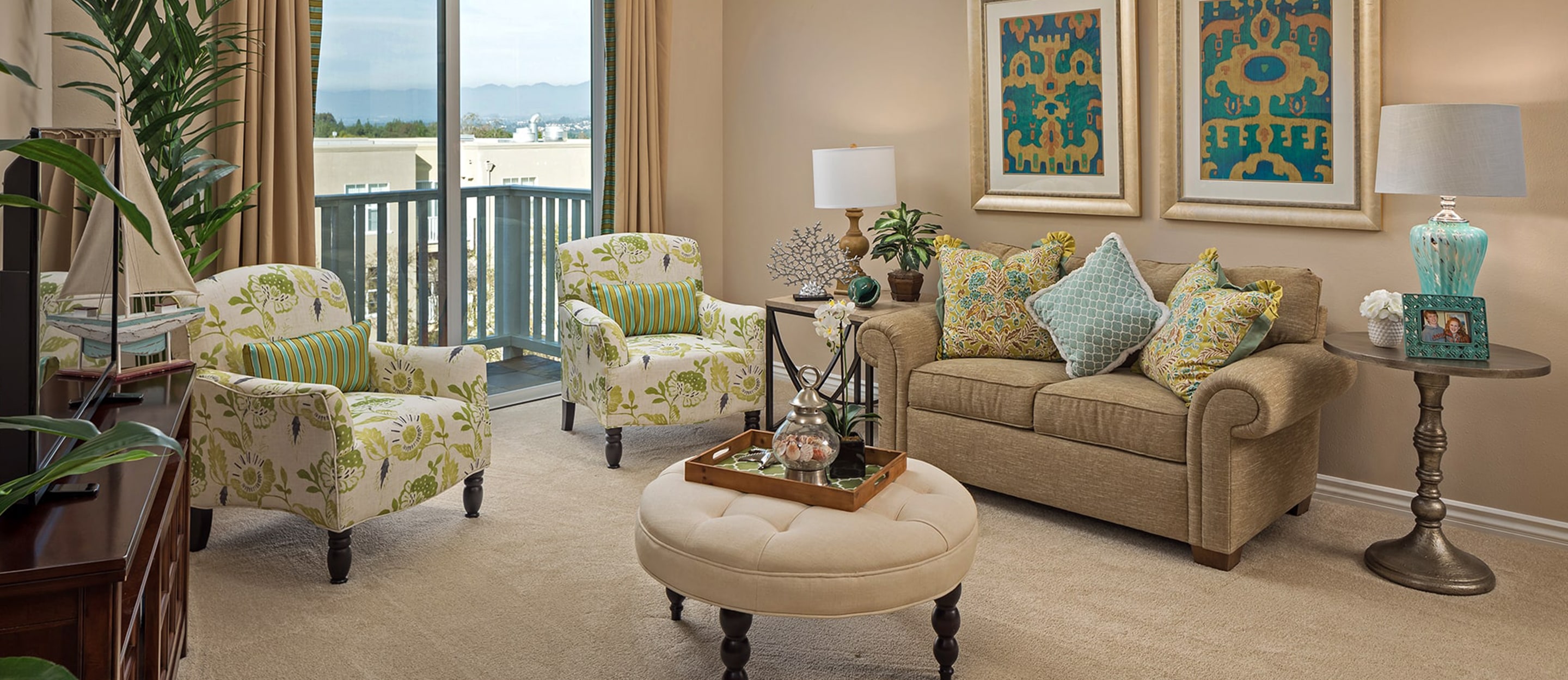 A cozy living room with a tan sofa, patterned armchairs, a round ottoman, and decorative pillows. Wall art, a table lamp, and a tray with candles are present. Sunlight streams in from large windows with a balcony view.