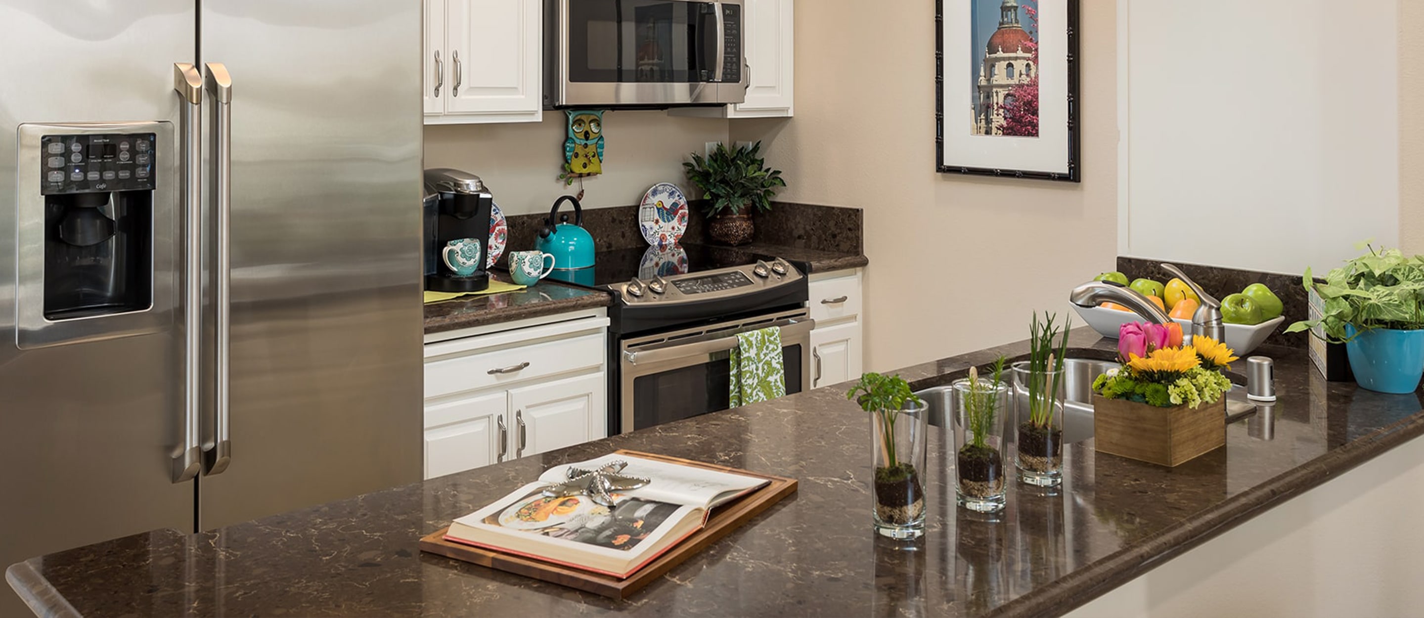 Modern kitchen with stainless steel fridge, black and brown countertops, white cabinets, microwave, stove, colorful dishes, open cookbook, three small plant glasses, and a box of flowers beside a sink with fresh fruits and vegetables.